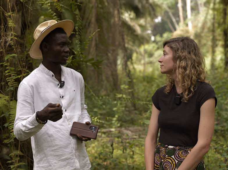 A man wearing a white shirt and a straw hat talks while holding a smartphone, facing a woman with curly hair wearing a black shirt and a patterned skirt, standing outdoors in a lush green forest.
