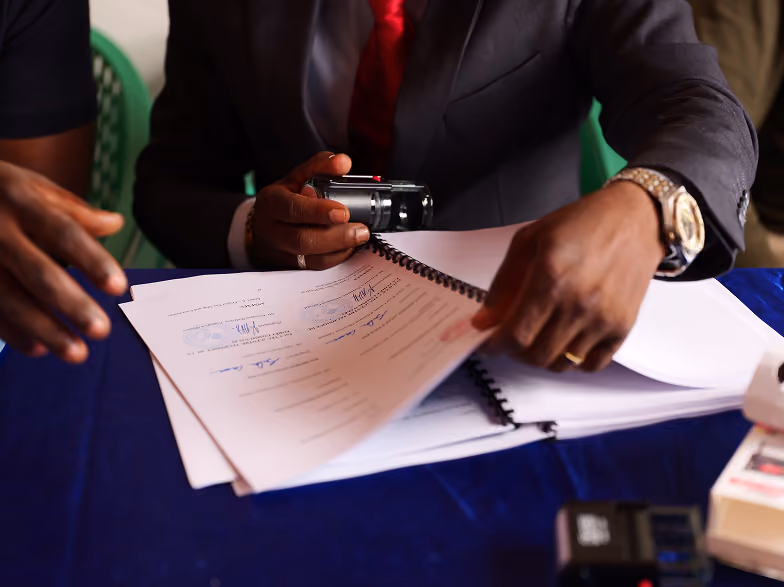 Close-up of a person in a suit and red tie holding a black stapler and flipping through spiral-bound documents with stamps and signatures on a blue tablecloth.