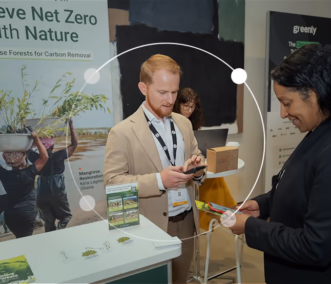 Two people at an informational booth about nature-based carbon removal; a man in a beige suit is using his phone while a woman in black examines a pamphlet, with posters and plant models in the background.