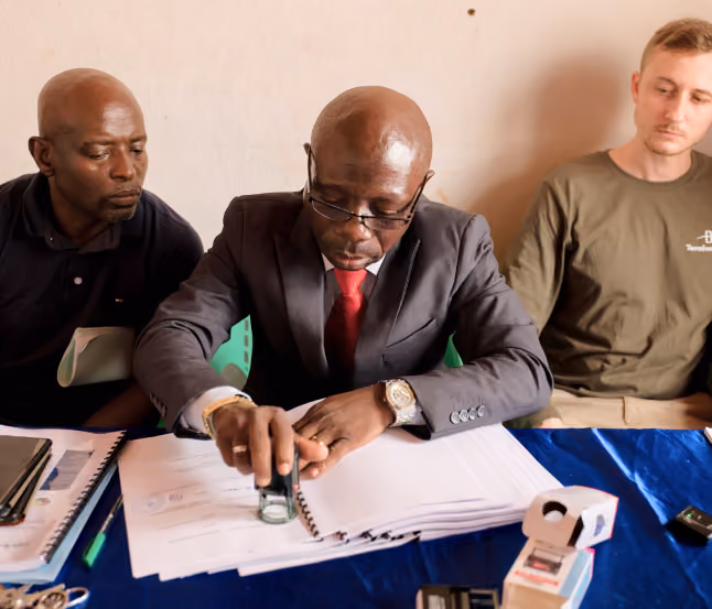Three men sitting at a table reviewing documents, the man in the center, wearing a suit and red tie, uses a stamp on papers while the others observe.