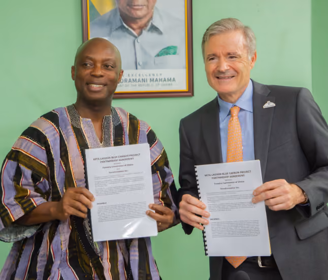 Two men smiling and holding signed documents labeled 'NITA-LAAGOON BLUE CARBON PROJECT PIPELINE AGREEMENT' in front of a green wall with a framed portrait of a man behind them.