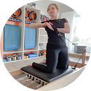 Woman kneeling on a Pilates reformer machine exercising with a resistance band in a bright studio.