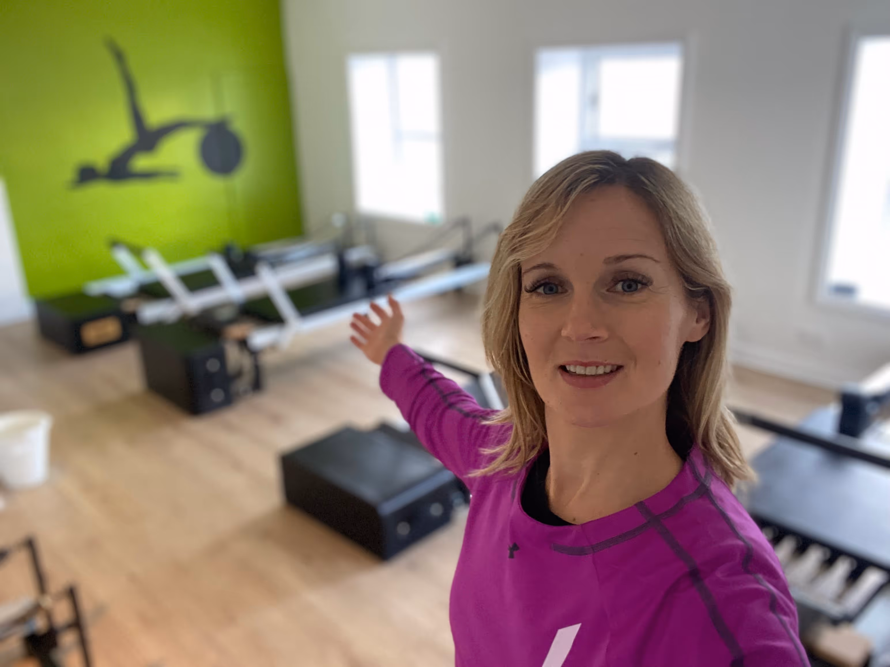 Woman in purple athletic shirt smiling and gesturing to empty exercise equipment in bright studio with green accent wall.