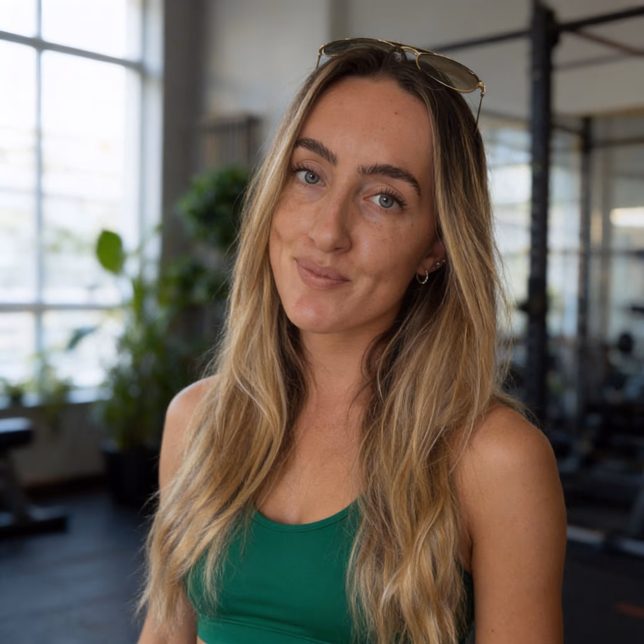 Young woman with long blonde hair wearing green workout top and sunglasses on her head in a gym setting.
