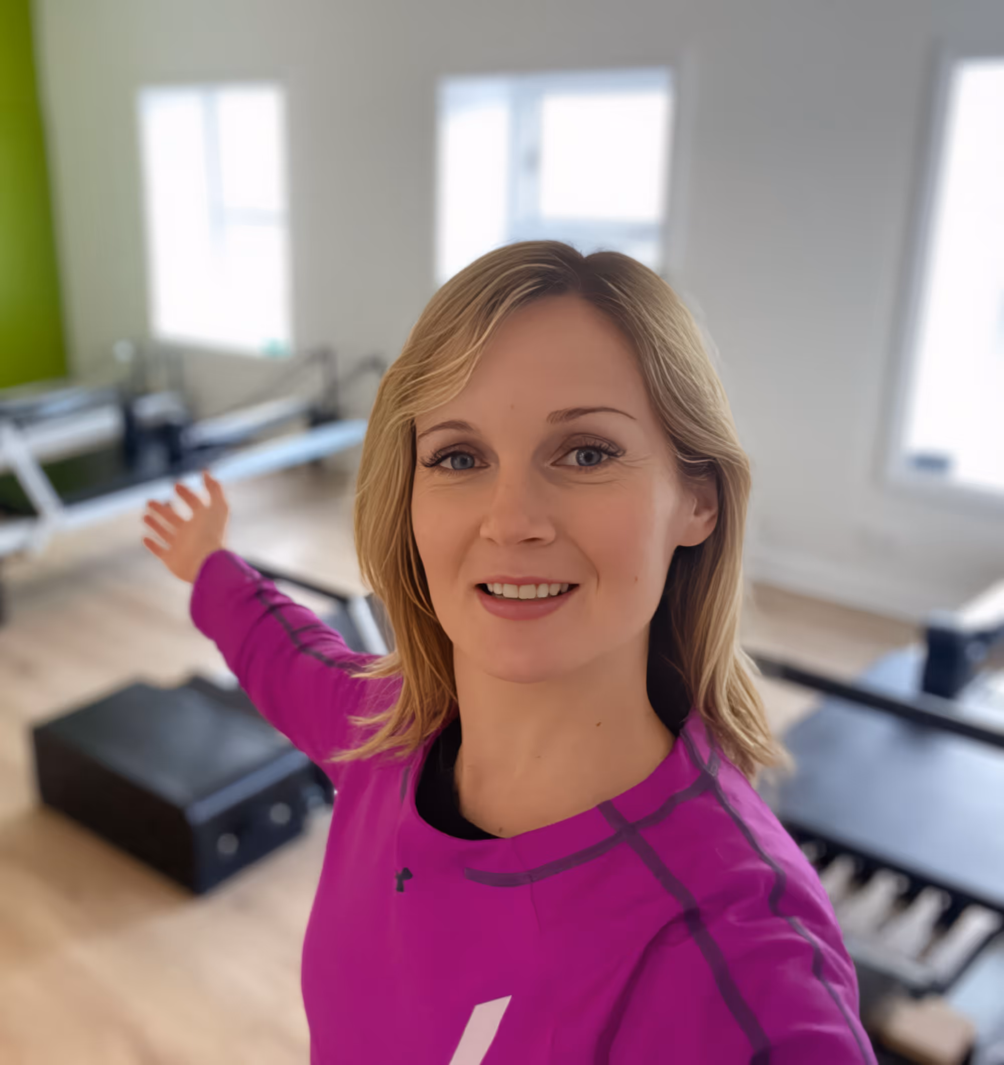 Smiling woman with blonde hair wearing a purple long-sleeve shirt, gesturing with her arm in a bright room with exercise equipment.