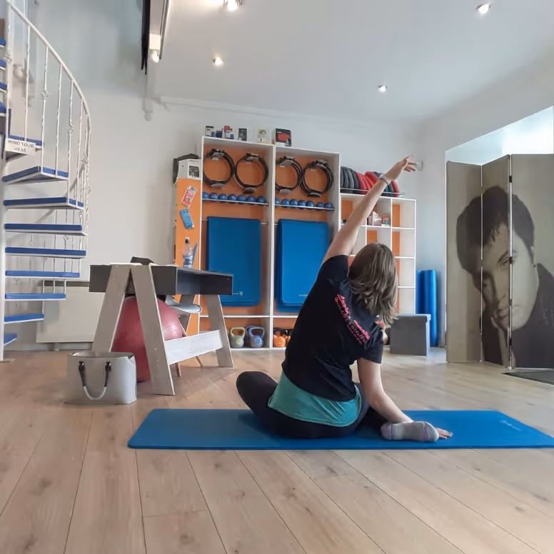 Person sitting on a blue exercise mat stretching arm upwards in a bright fitness room with gym equipment and a large portrait on a folding screen.