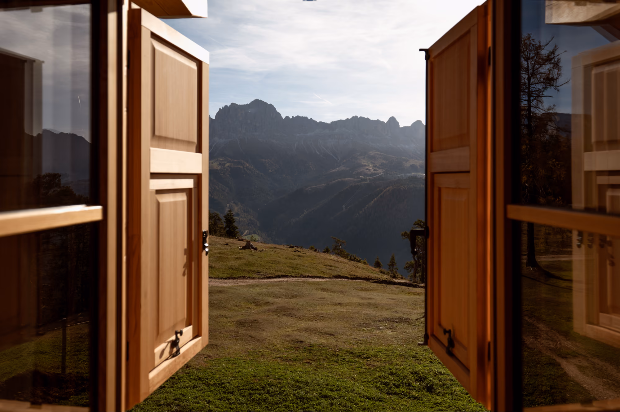 View of mountain range seen through open wooden window shutters with grassy hill in foreground.