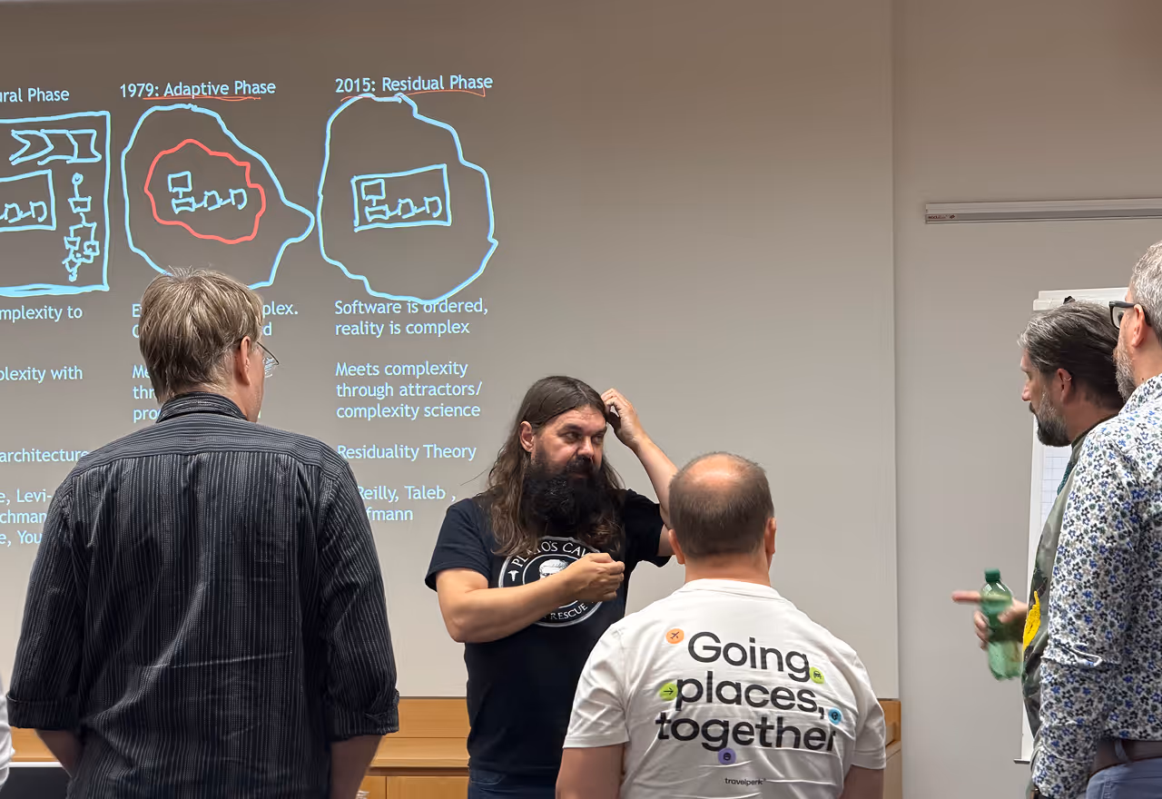 Five men stand discussing a presentation slide with diagrams and text about software complexity phases.