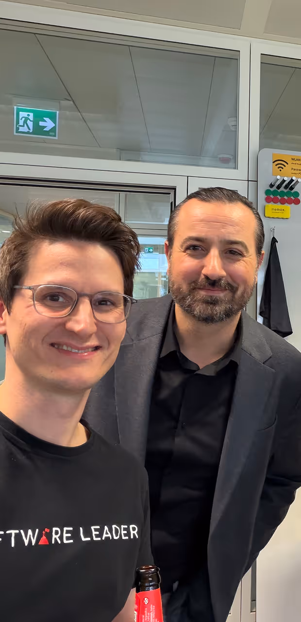 Two men smiling indoors; one wearing glasses and a Software Leader shirt holding a bottle.