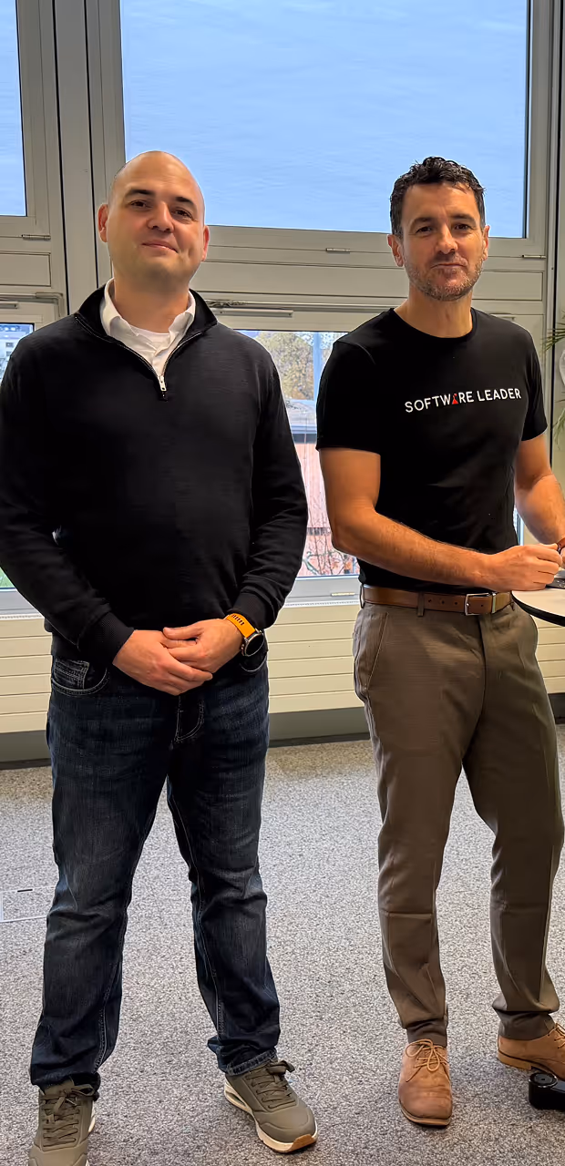 Two men standing indoors; one wears a black pullover, the other a black 'Software Leader' t-shirt.