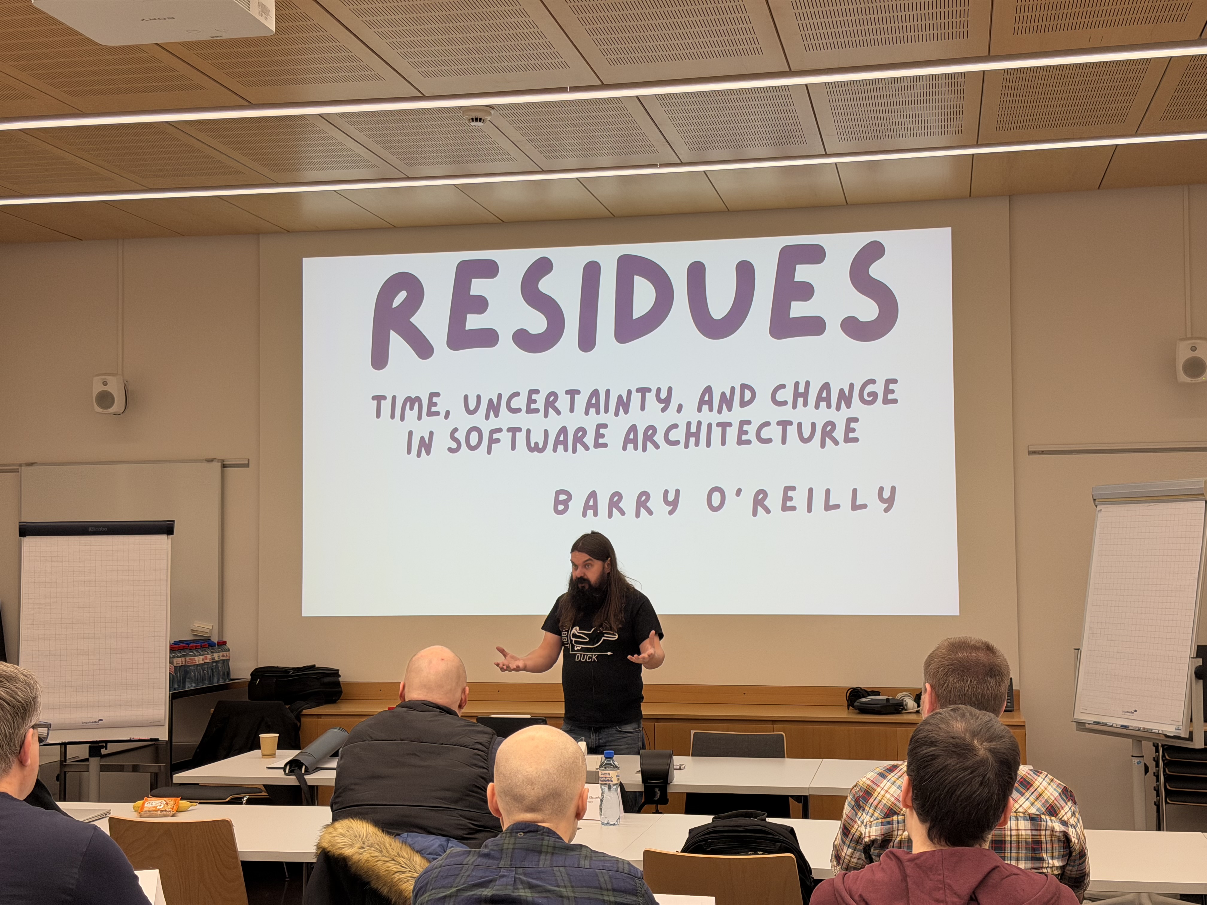 Man with long hair giving a talk titled 'RESIDUES Time, Uncertainty, and Change in Software Architecture' by Barry O'Reilly.