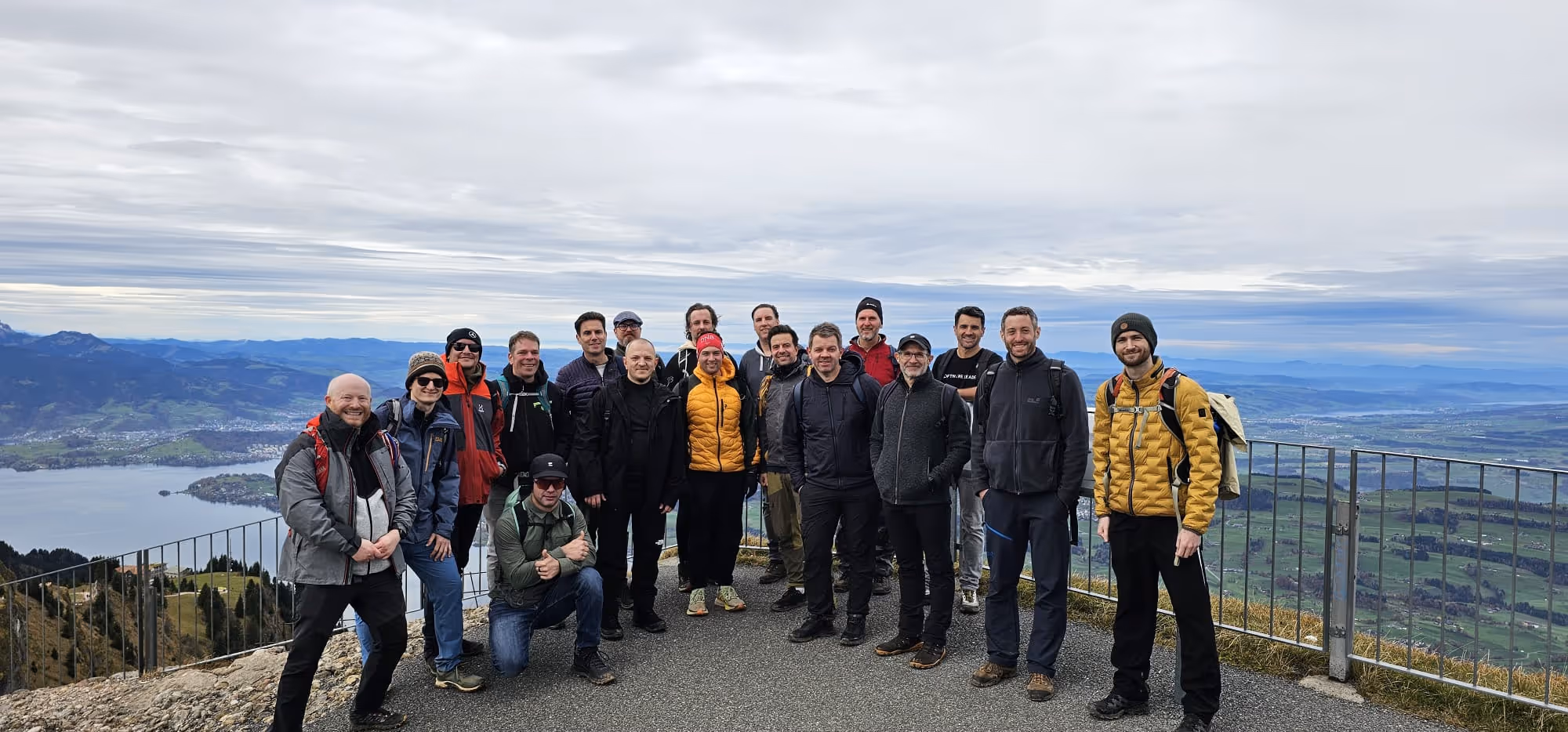 Group of sixteen hikers posing on a mountain overlook with a lake and valley view behind them.