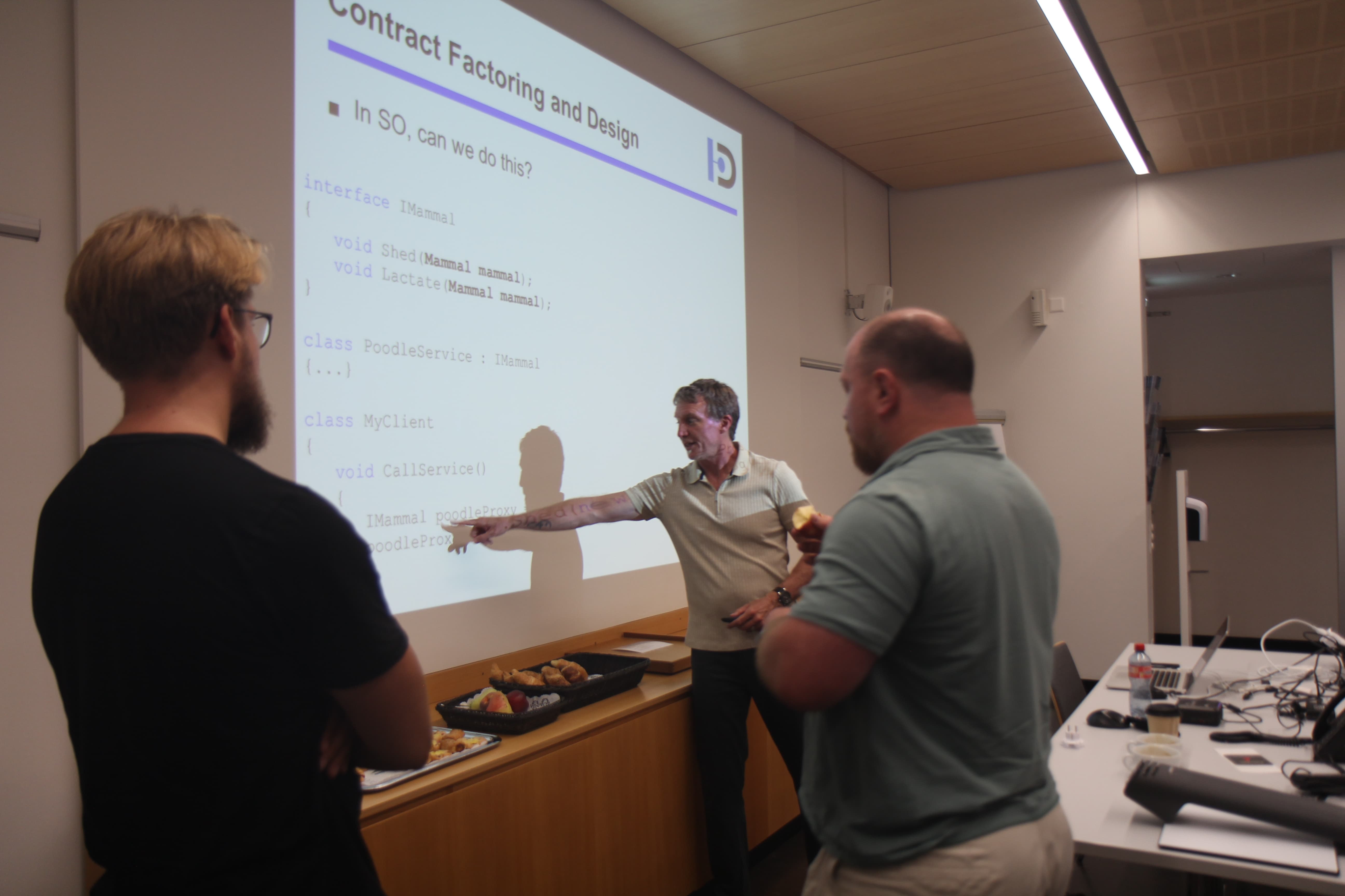 Man points at projected slide with code while two men listen in a meeting room with snacks and laptops.