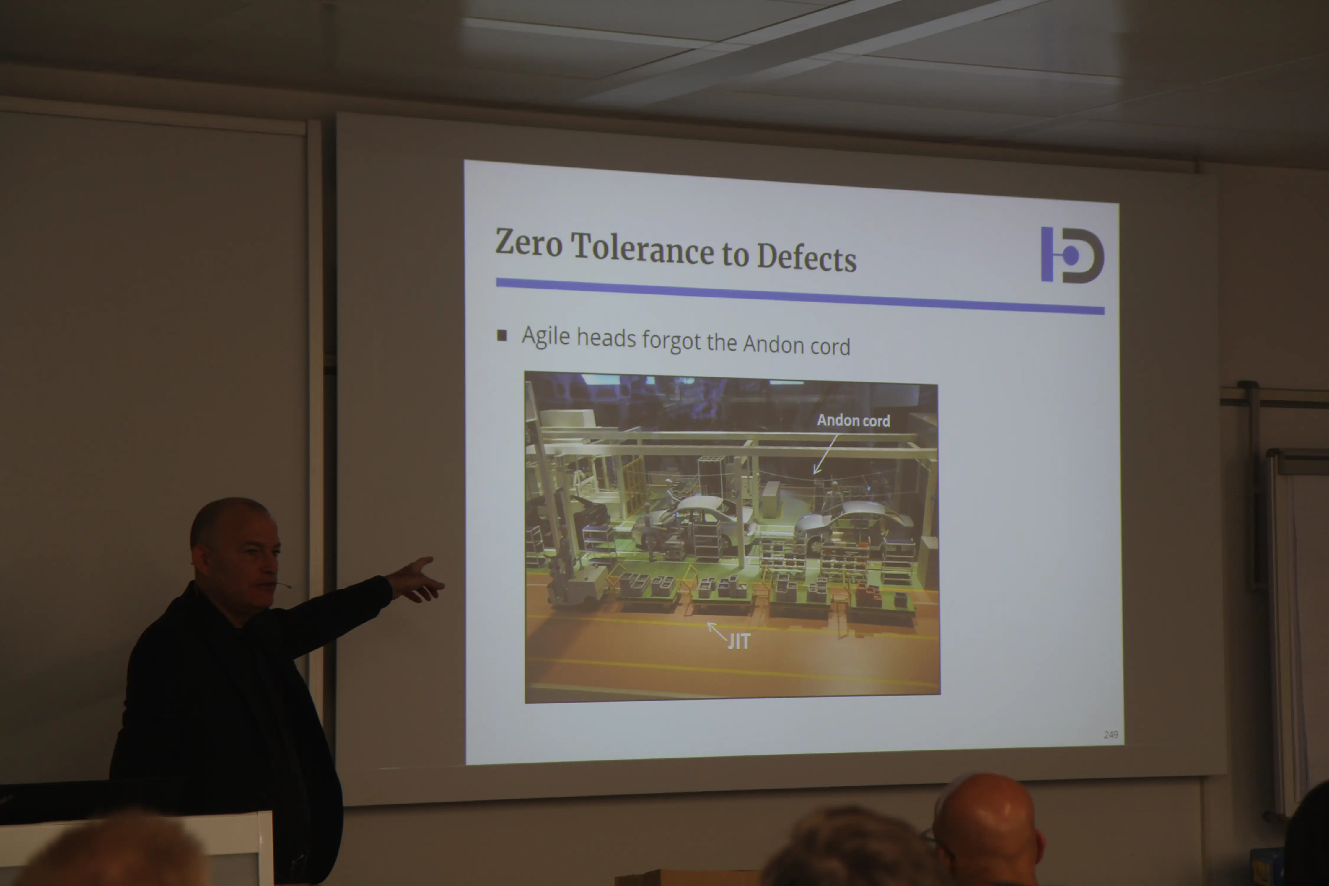 Presenter pointing to slide titled 'Zero Tolerance to Defects' discussing Agile and Andon cord in manufacturing.