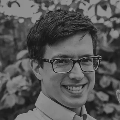 Smiling man with short dark hair and glasses, blurred foliage background.