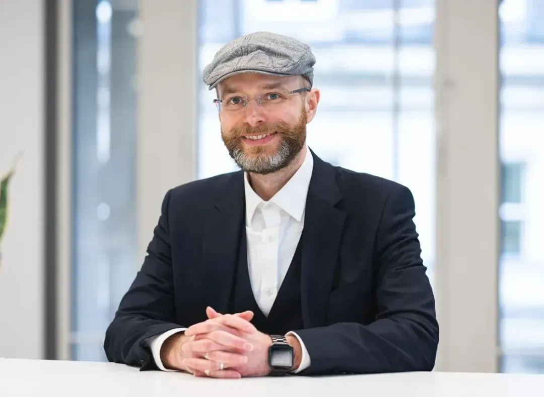 Bearded man wearing glasses, a gray flat cap, and a black suit sitting at a table with hands clasped.