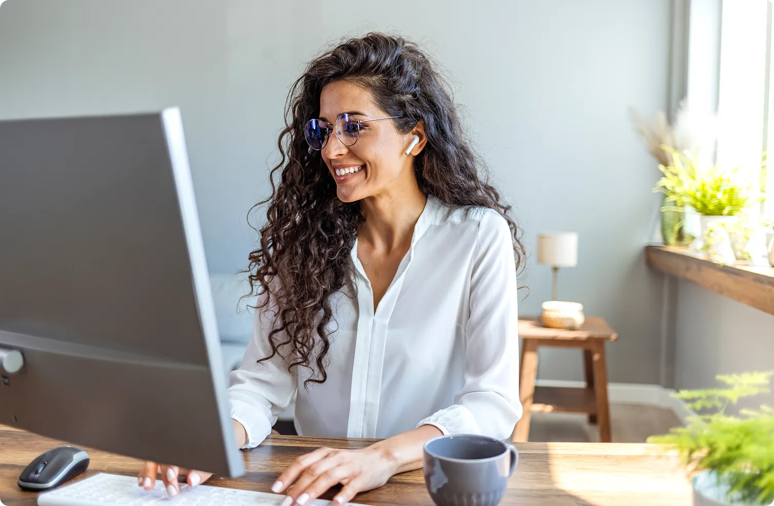 Smiling woman with curly hair and glasses working on a desktop computer at a sunny wooden desk.
