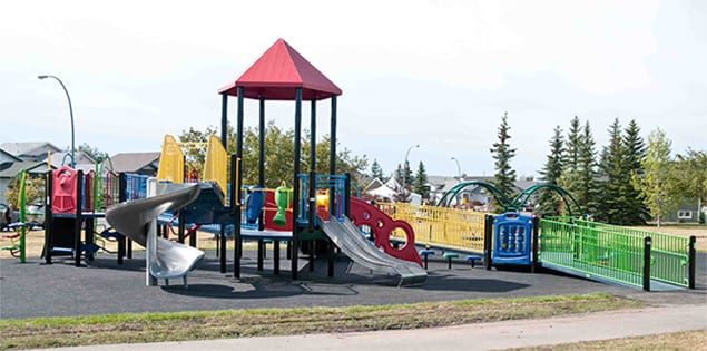 Wide view of colorful playground with red-peaked tower, spiral and straight slides, ramps and green perimeter fence.