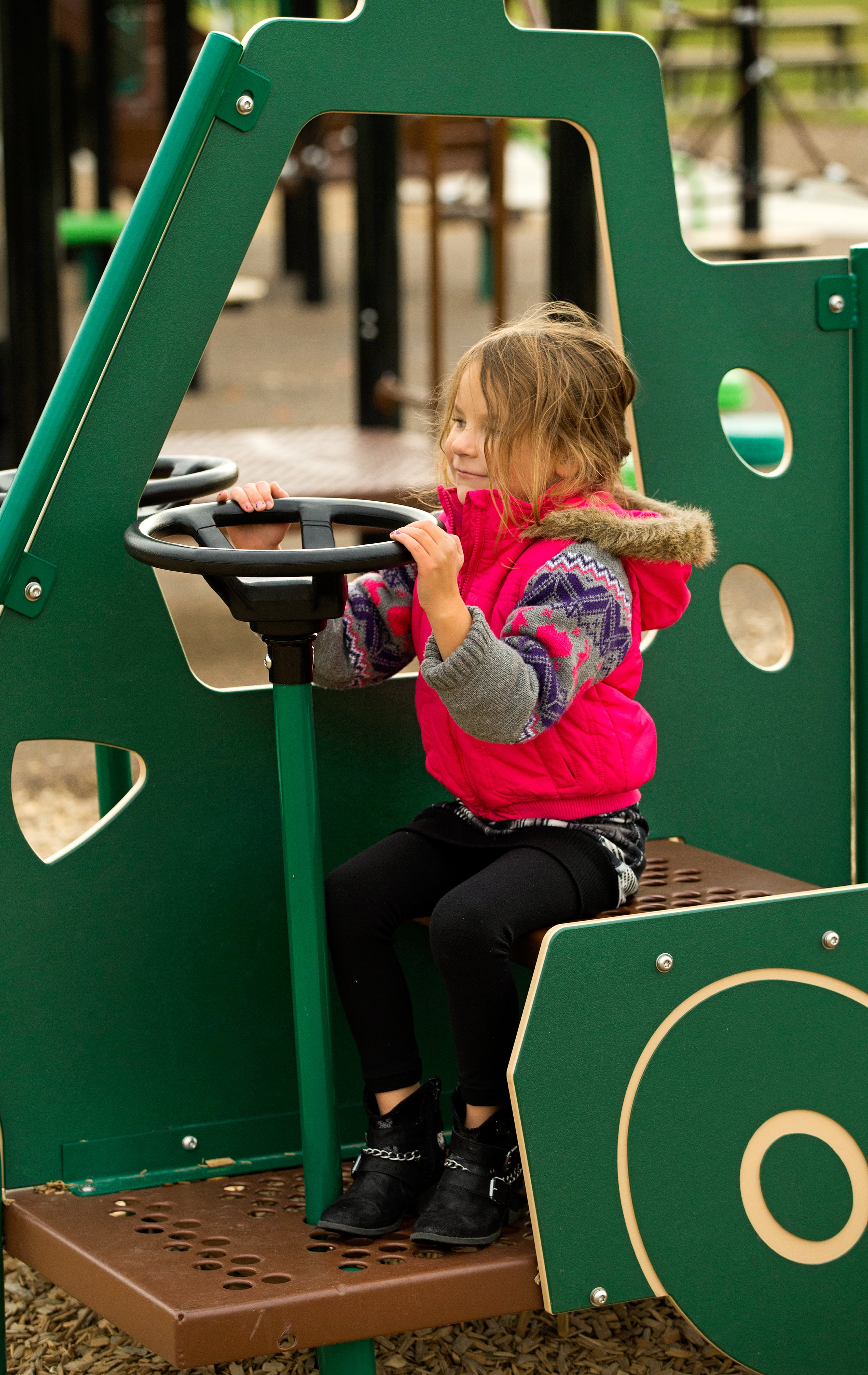 Child wearing a pink vest grips a black steering wheel while seated inside a green play vehicle; close view of hands and face.