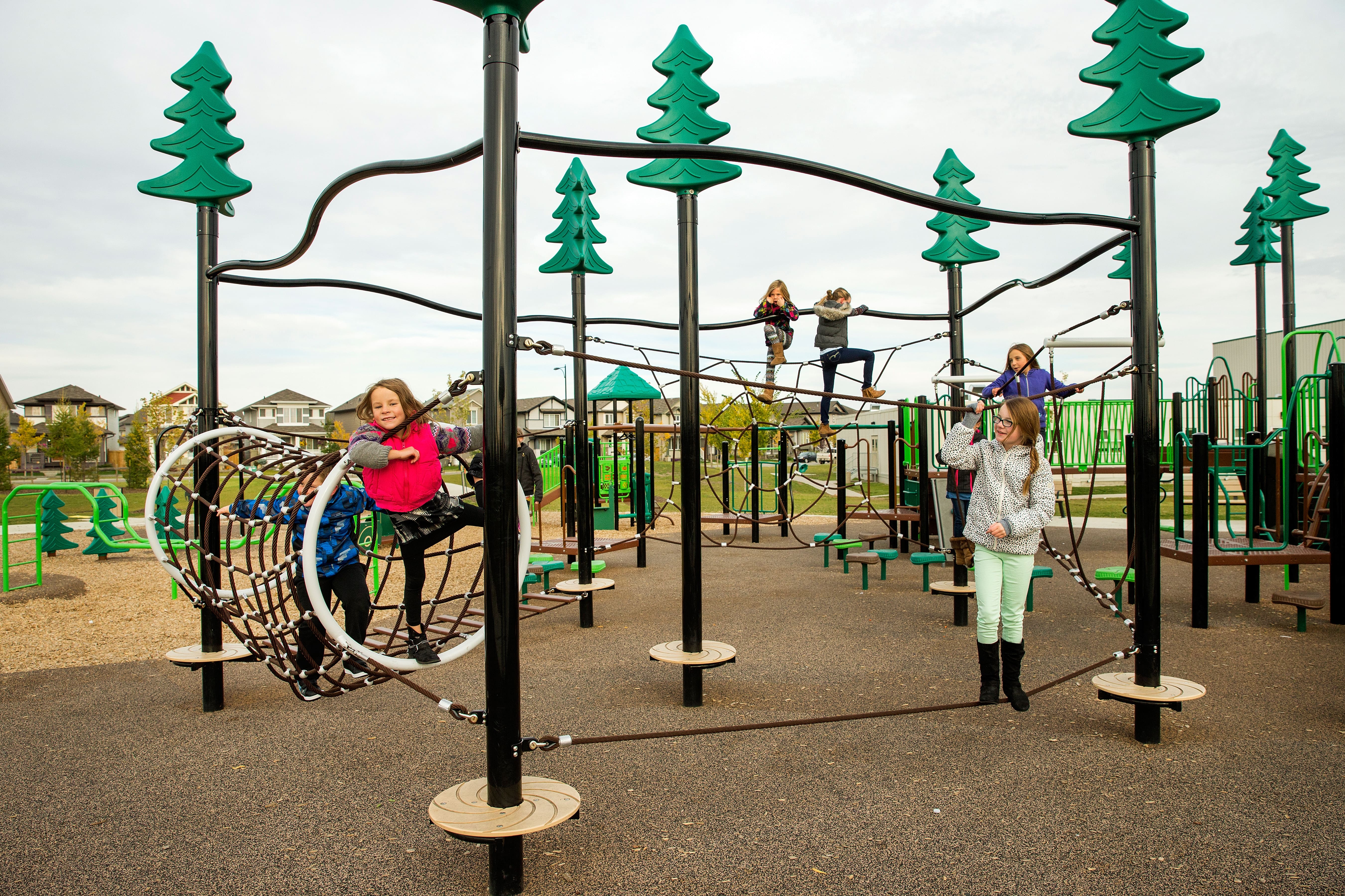Children climbing and playing on a black-and-green rope obstacle course with a circular rope tunnel and stylized green tree-top posts.
