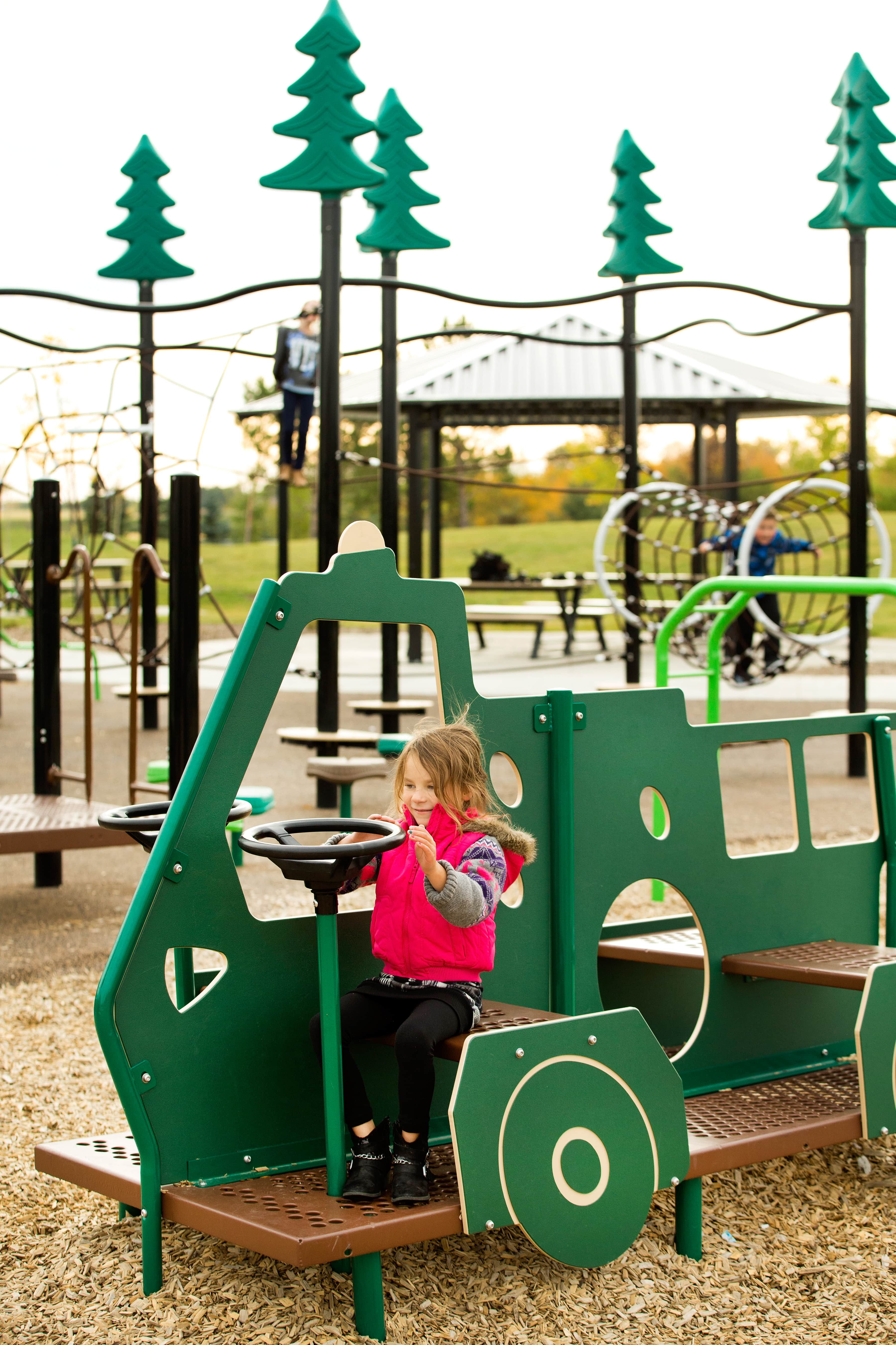 Child at the steering wheel of a green play-vehicle panel, wood-chip surfacing visible and rope-climb features and shelter in background.