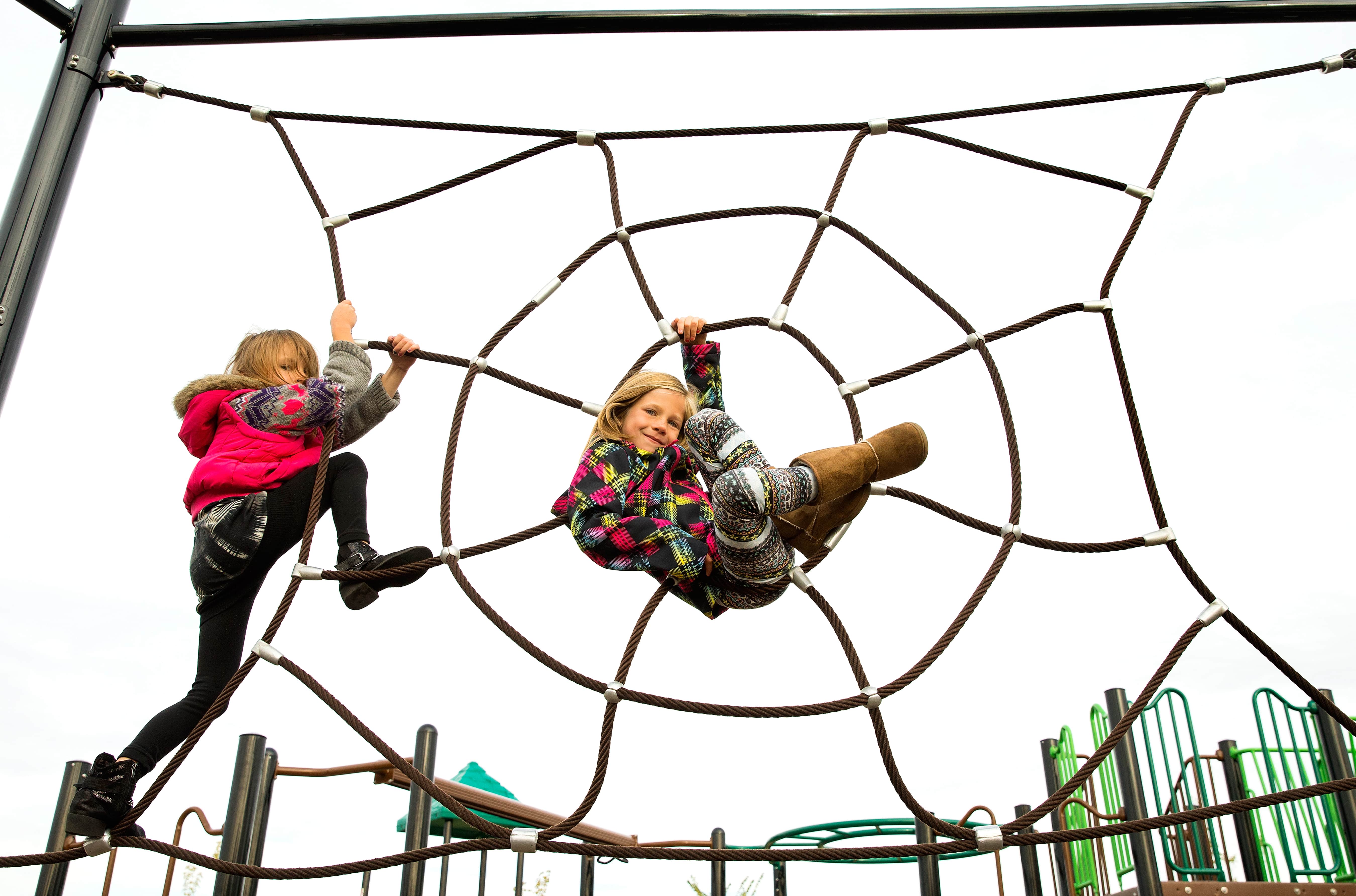 Two girls climbing a large circular rope web; one sits in the center wearing patterned leggings and boots while another ascends at left.