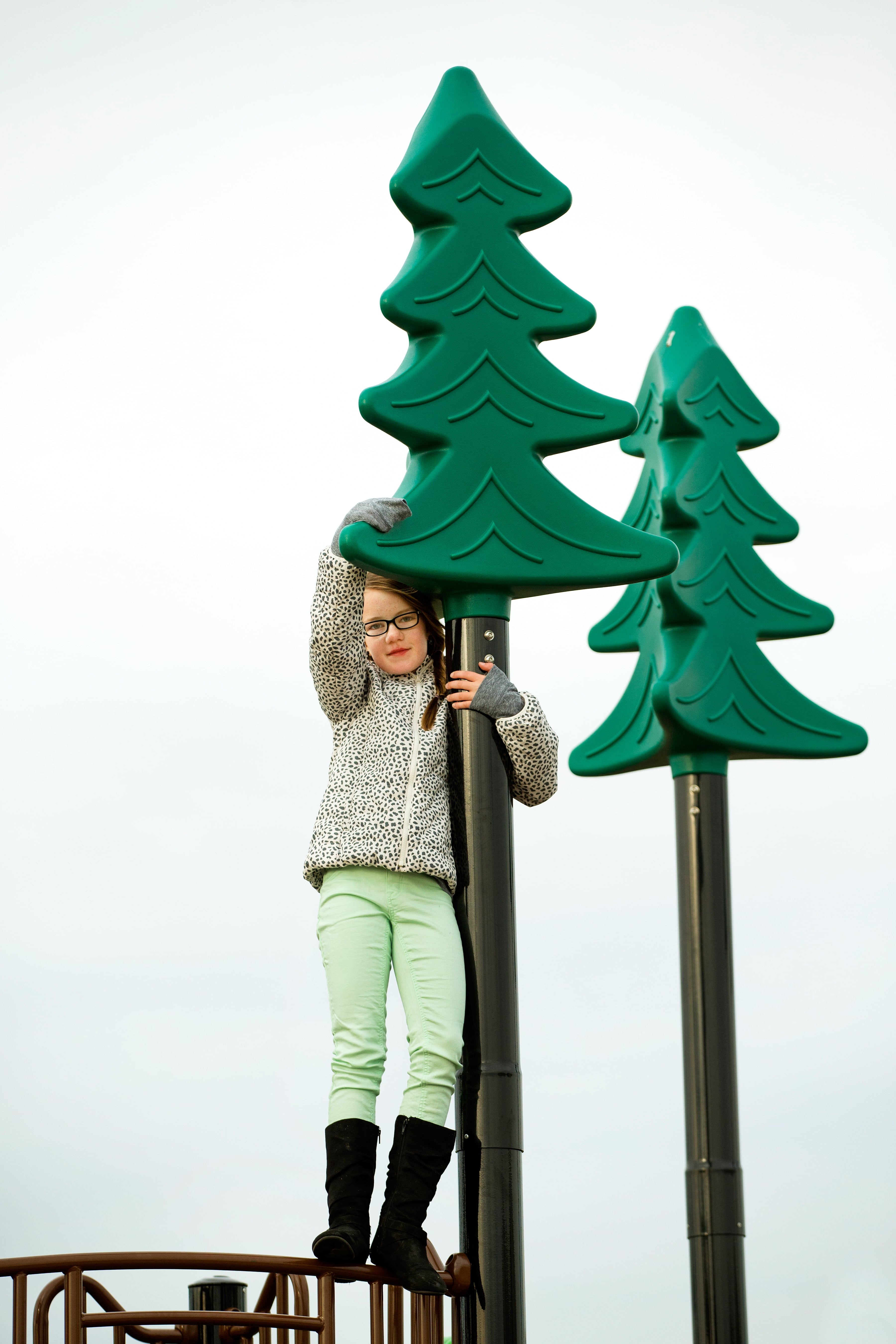 Child with glasses gripping a black pole topped by a green stylized tree, standing on a textured climbing platform.