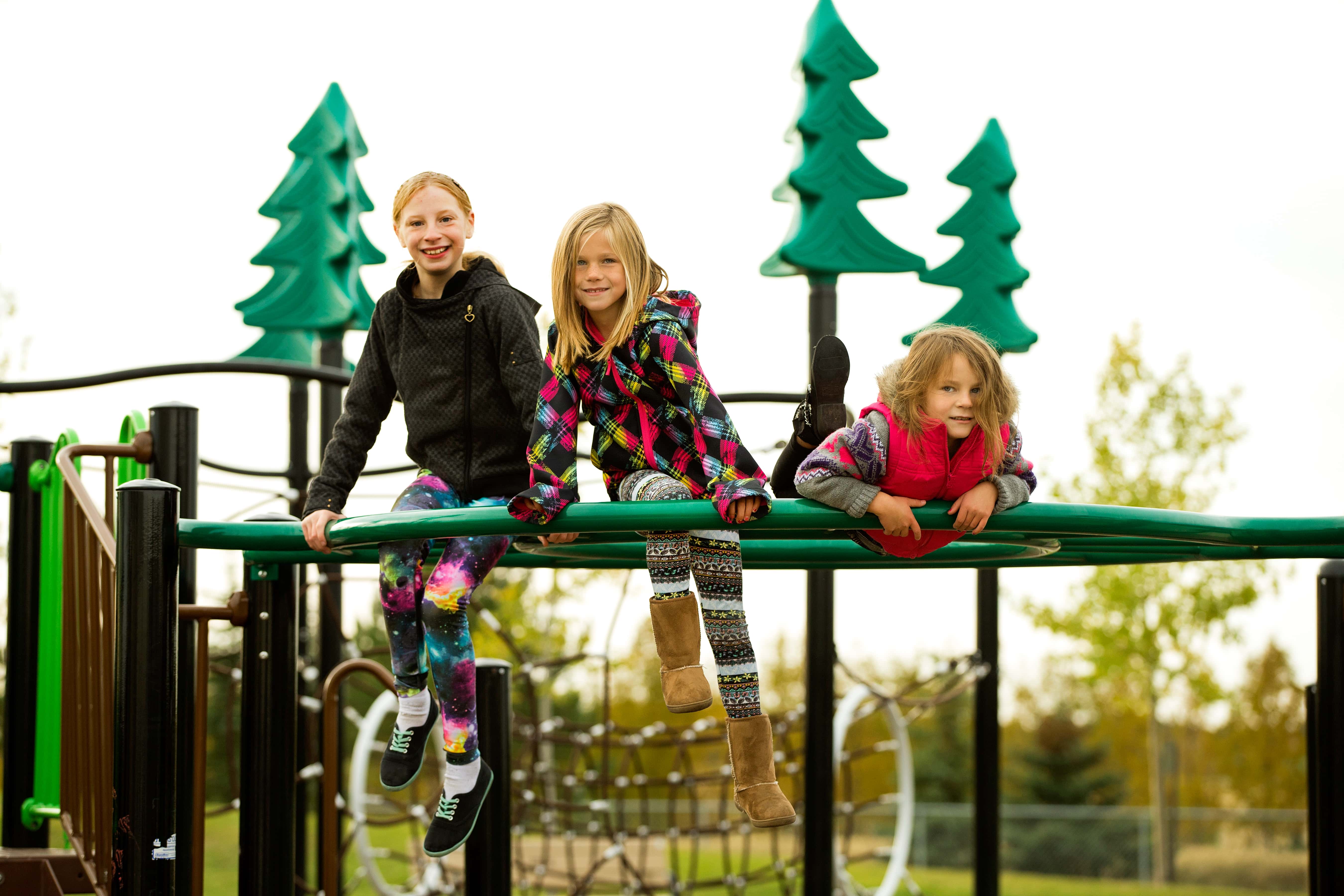 Three children perched on a curved green rail of the play structure — two sitting and one draped over the bar with trees behind.