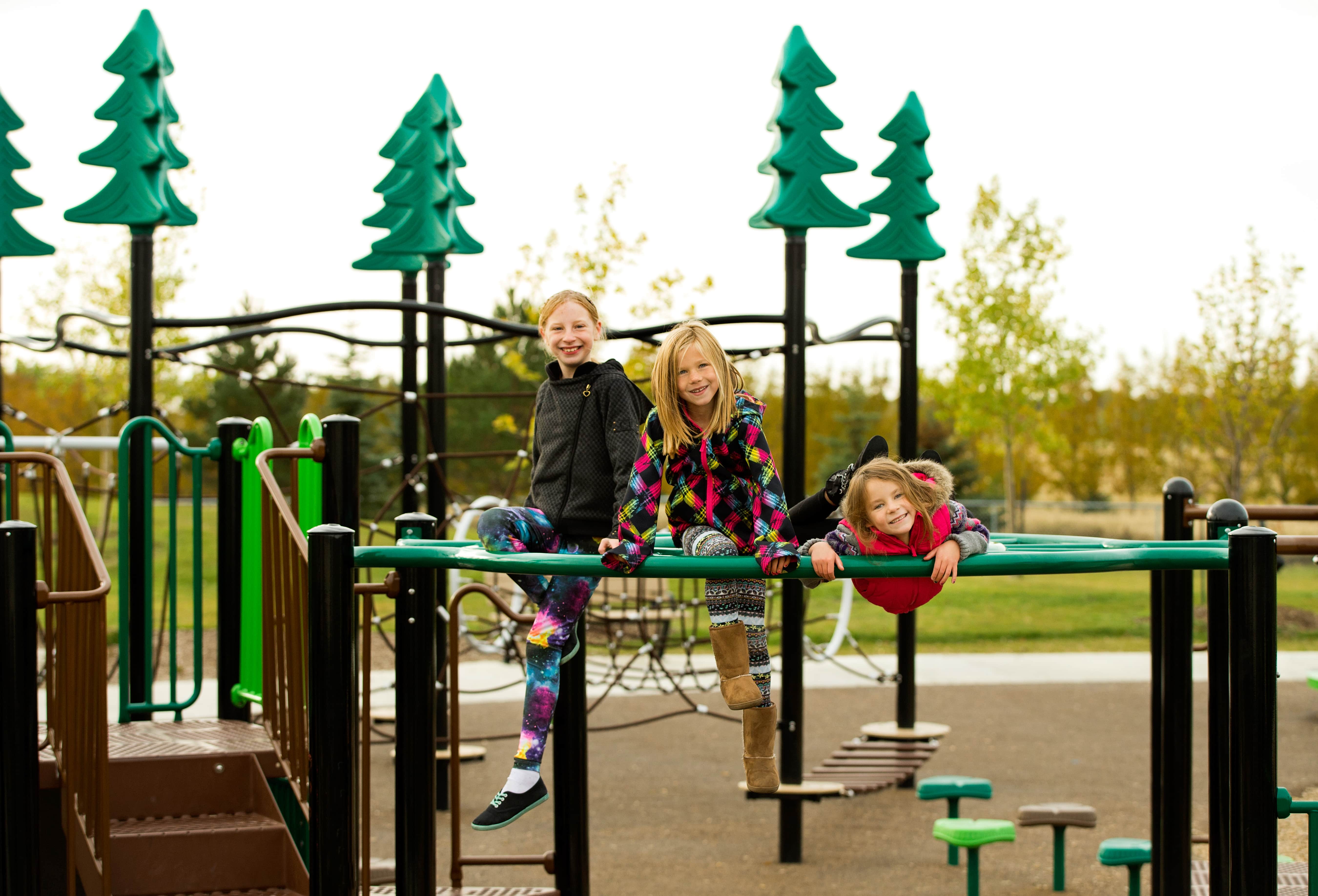 Three children sitting and lying on a green horizontal bar across the play structure, smiling with tree-topped poles behind.