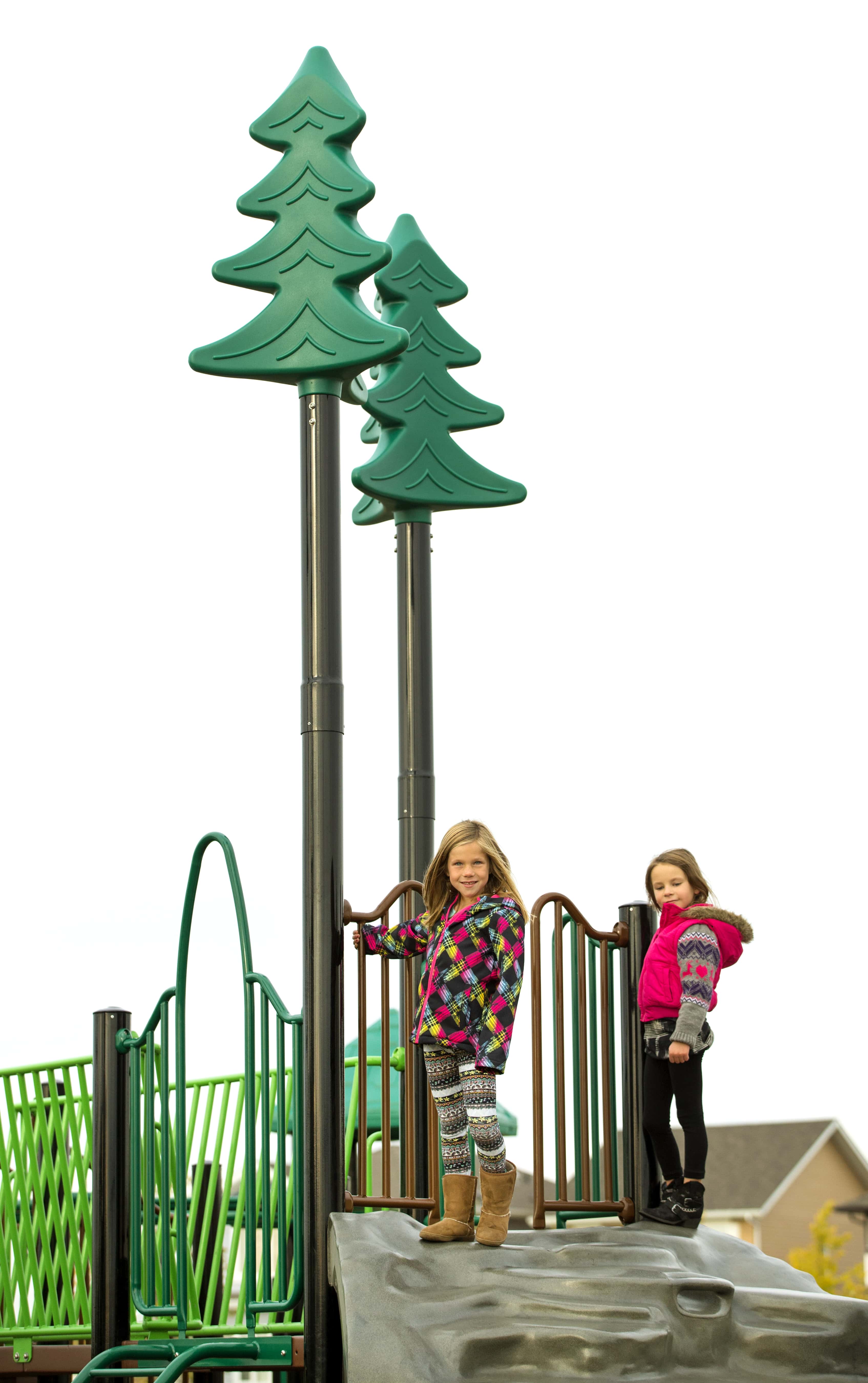 Two girls standing on a playground platform beneath tall poles topped with green tree-shaped caps, wearing colorful jackets.