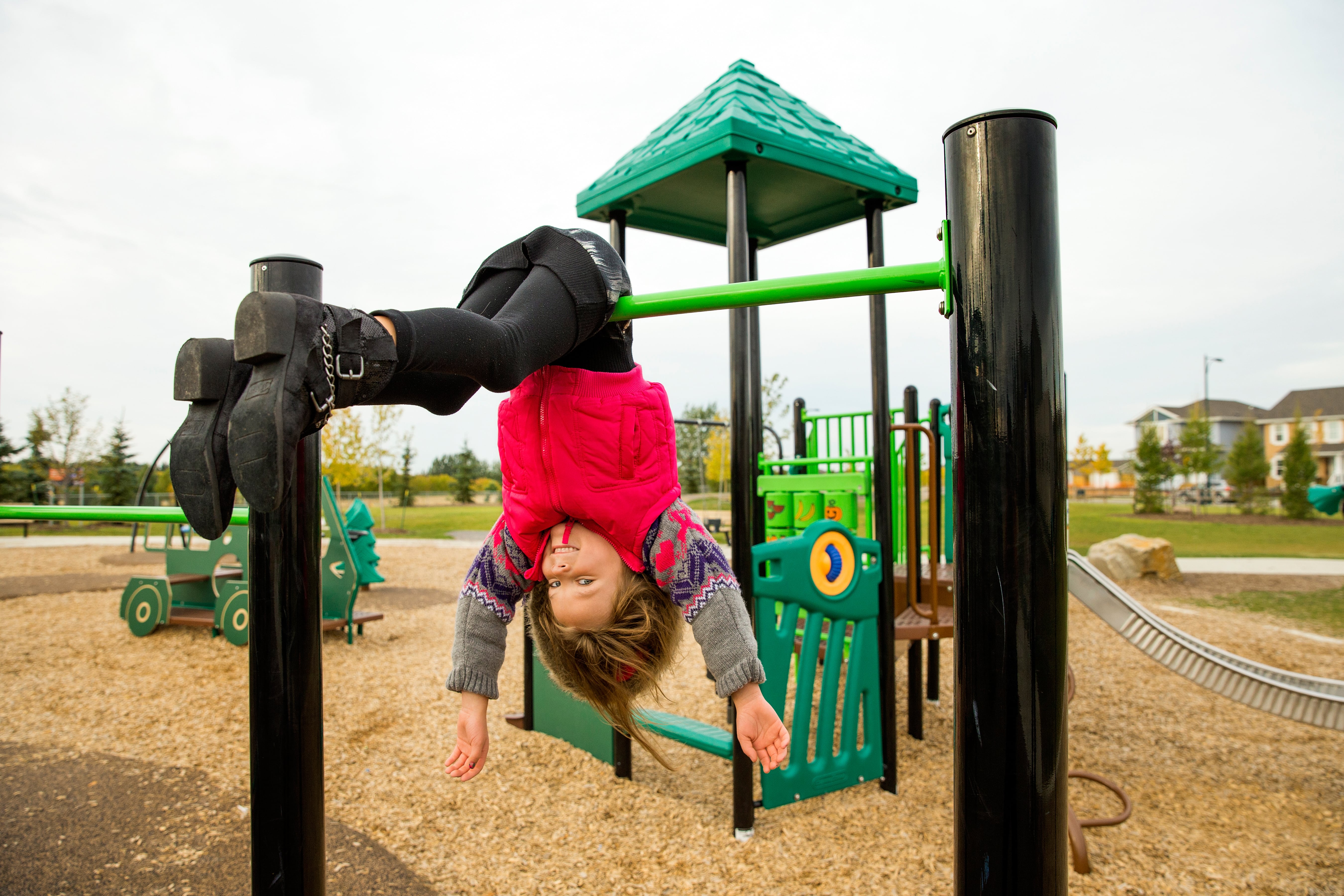 Child held upside down by another while hanging over a low green bar, with wood-chip surfacing and playground pieces visible around them.