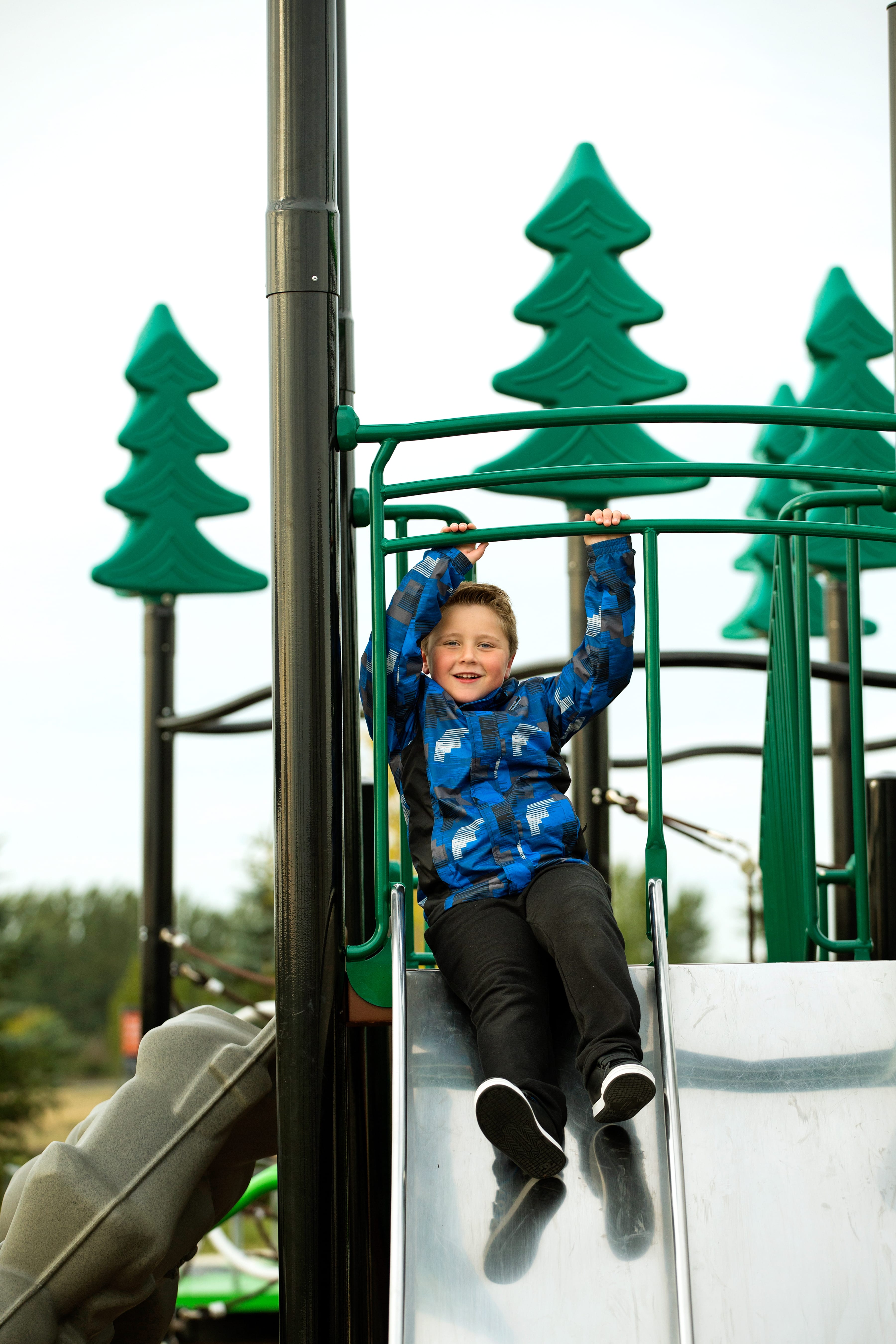 Boy smiling as he slides down a metal slide, hands gripping green safety rails with tree-shaped toppers visible above.