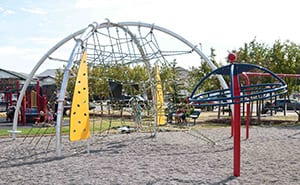 Dome-shaped metal climbing net with yellow climbing panels beside a curved blue-and-red track structure over gravel.