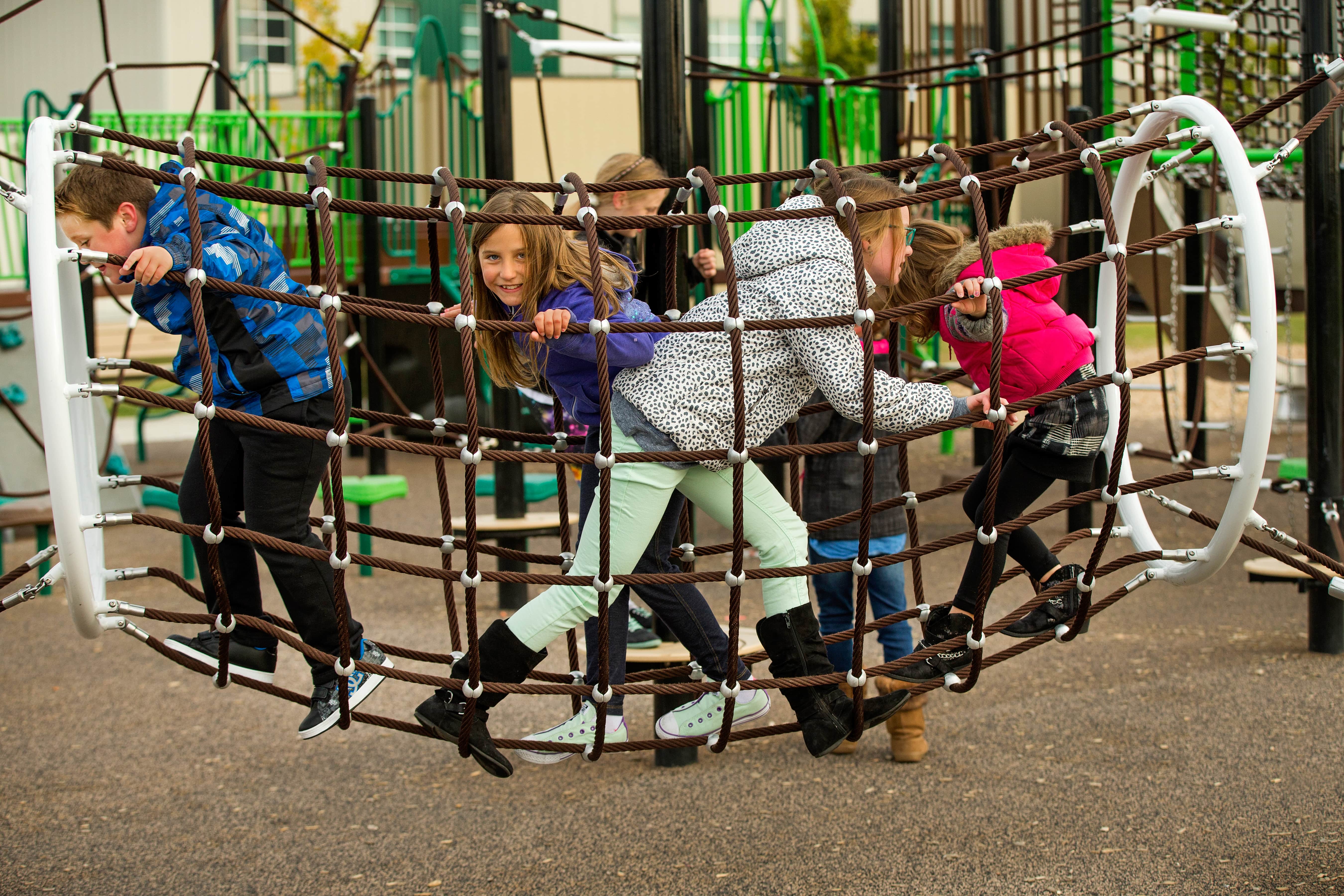 Several children crawl and climb inside a horizontal rope tunnel net, peering through square openings as they move across the web.