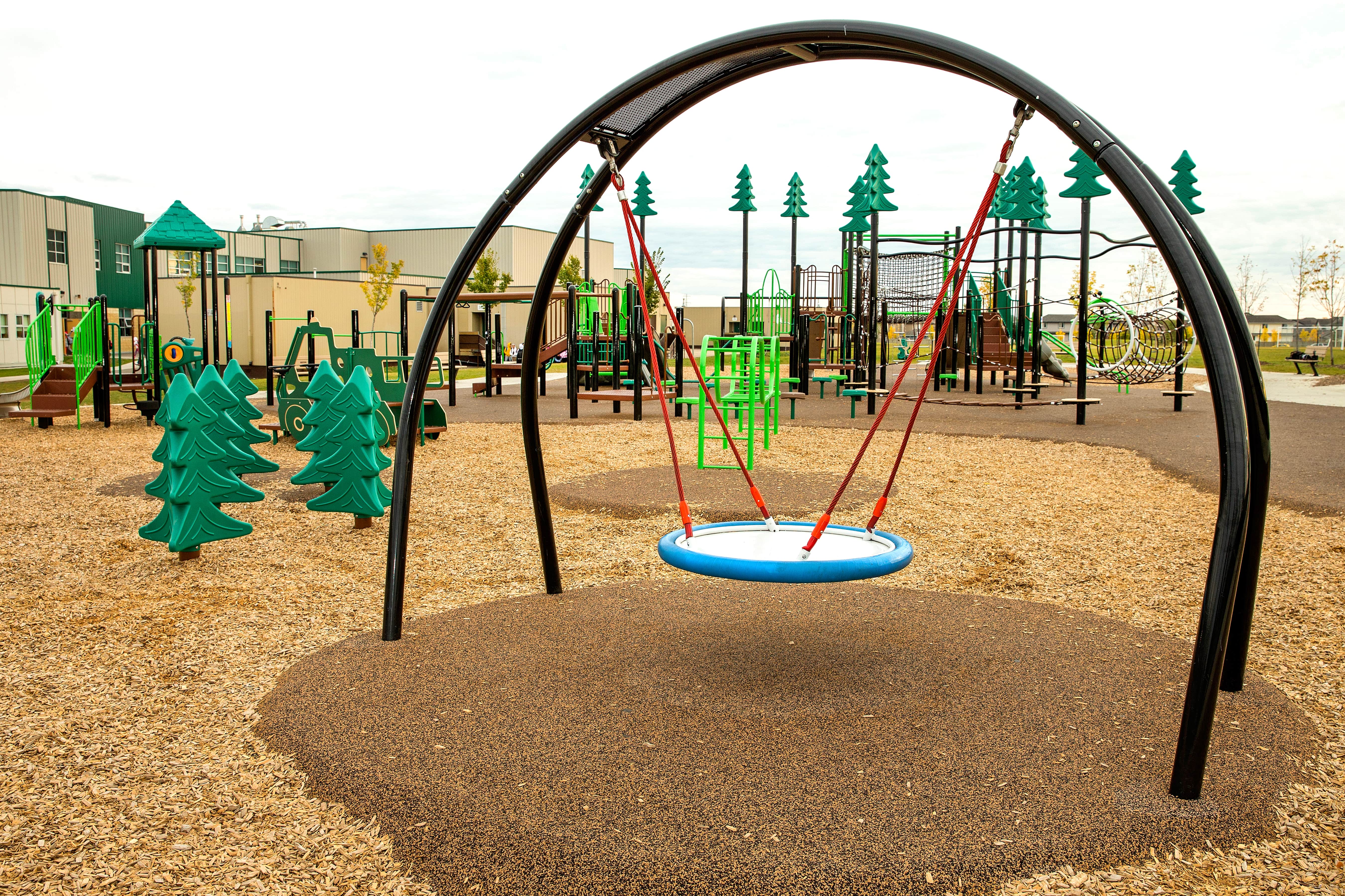 Blue circular nest swing hanging from a black arch, framed by green tree-shaped play panels with multi-level playground structures behind.