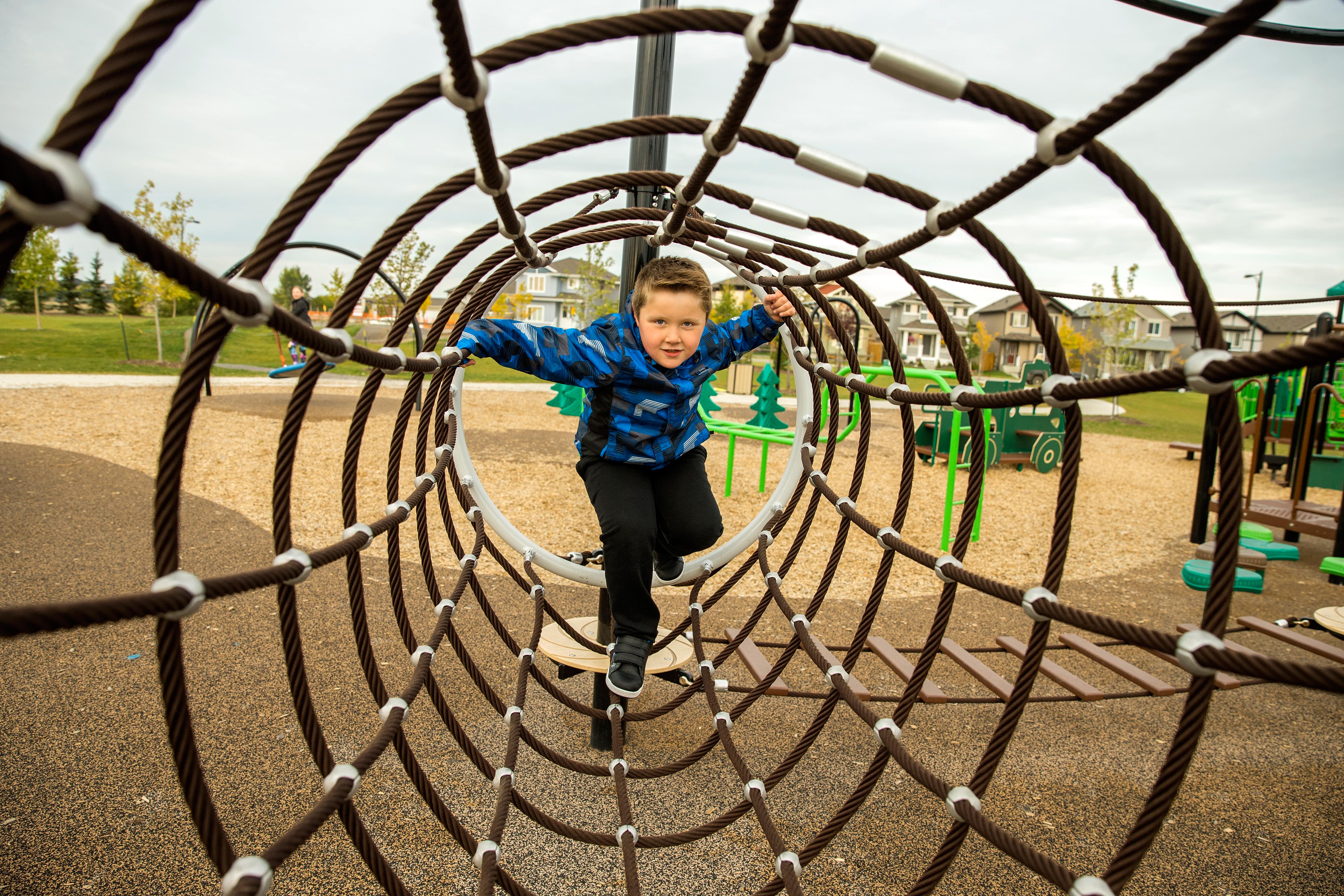 Boy crawls through concentric rope rings toward the camera, framed by circular netting with playground equipment visible beyond.