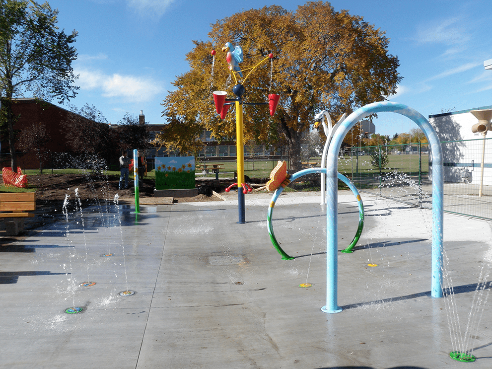 Concrete splash pad featuring light-blue arching spray rings and a tall yellow pole topped with a blue bird figure, autumn tree behind.