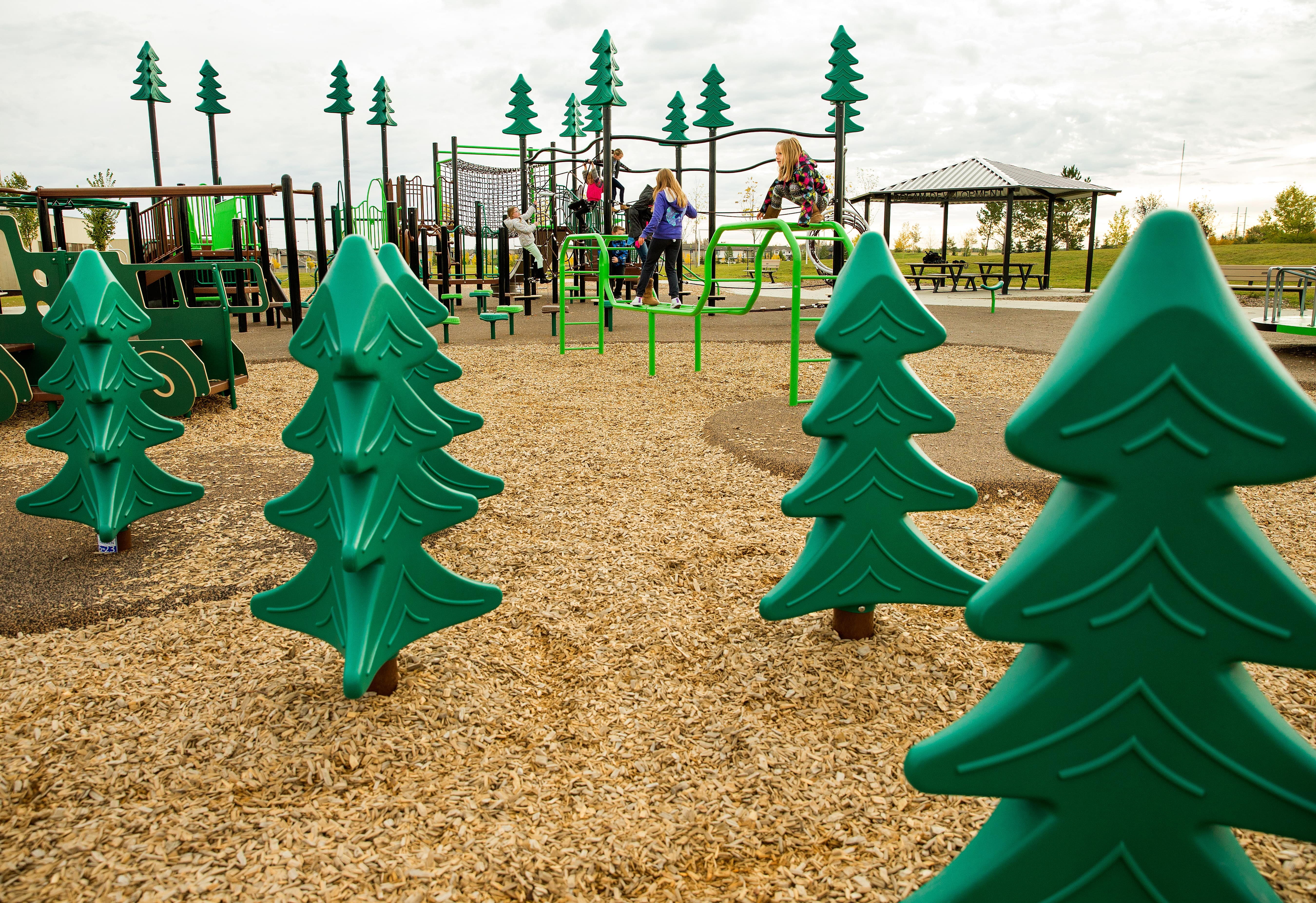 Row of green tree-shaped play panels in the foreground framing children climbing platforms, balance steps and a large rope obstacle course.
