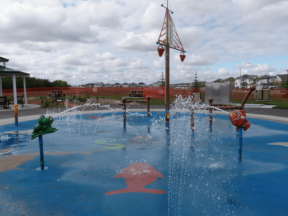 Blue splash pad surface with frog and fish spray features spouting water and a central pole topped by a triangular bucket element.