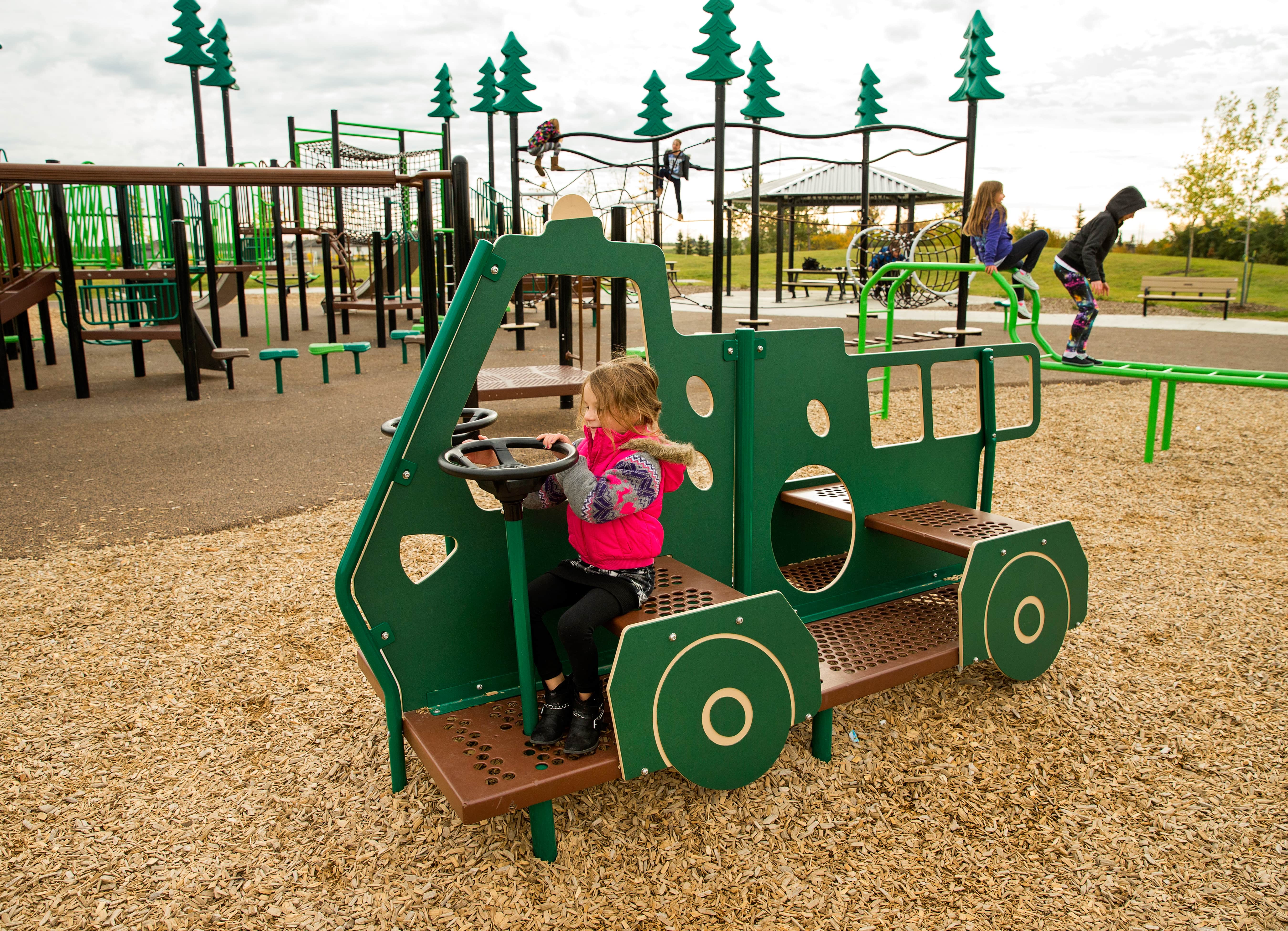 Green play-vehicle in the foreground with a child at the steering wheel; rope-climb structure, slides and swings visible across the playground.