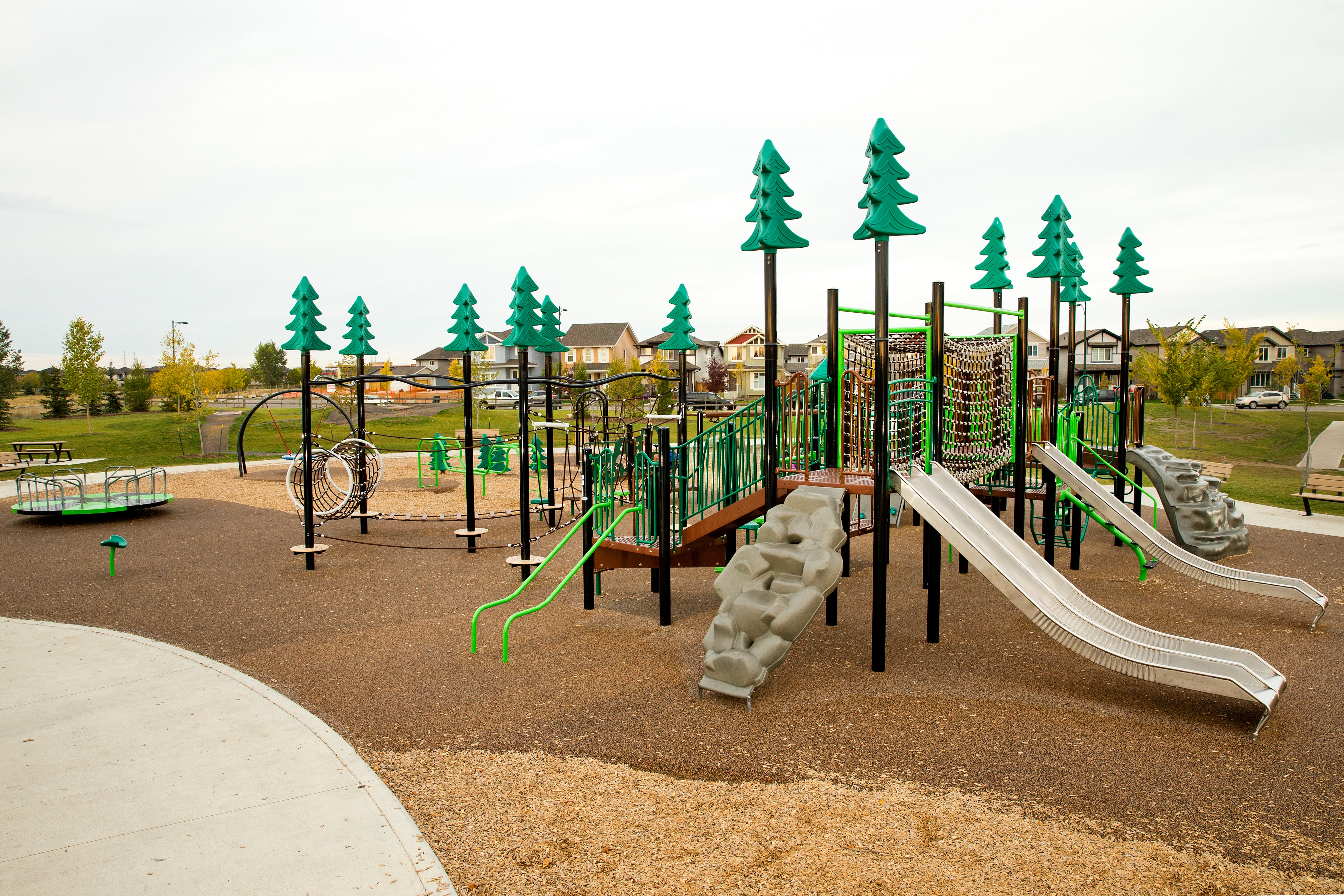 Wide empty view of the playground showing twin metal slides, rope-net climbing structures, a merry-go-round and multiple green tree-top posts.