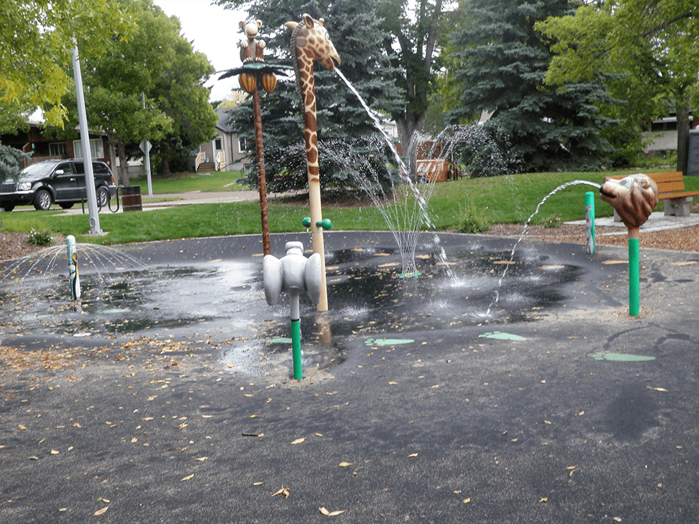 Play splash pad with a tall giraffe head spraying water across a wet black surface, animal spray heads and trees beyond.