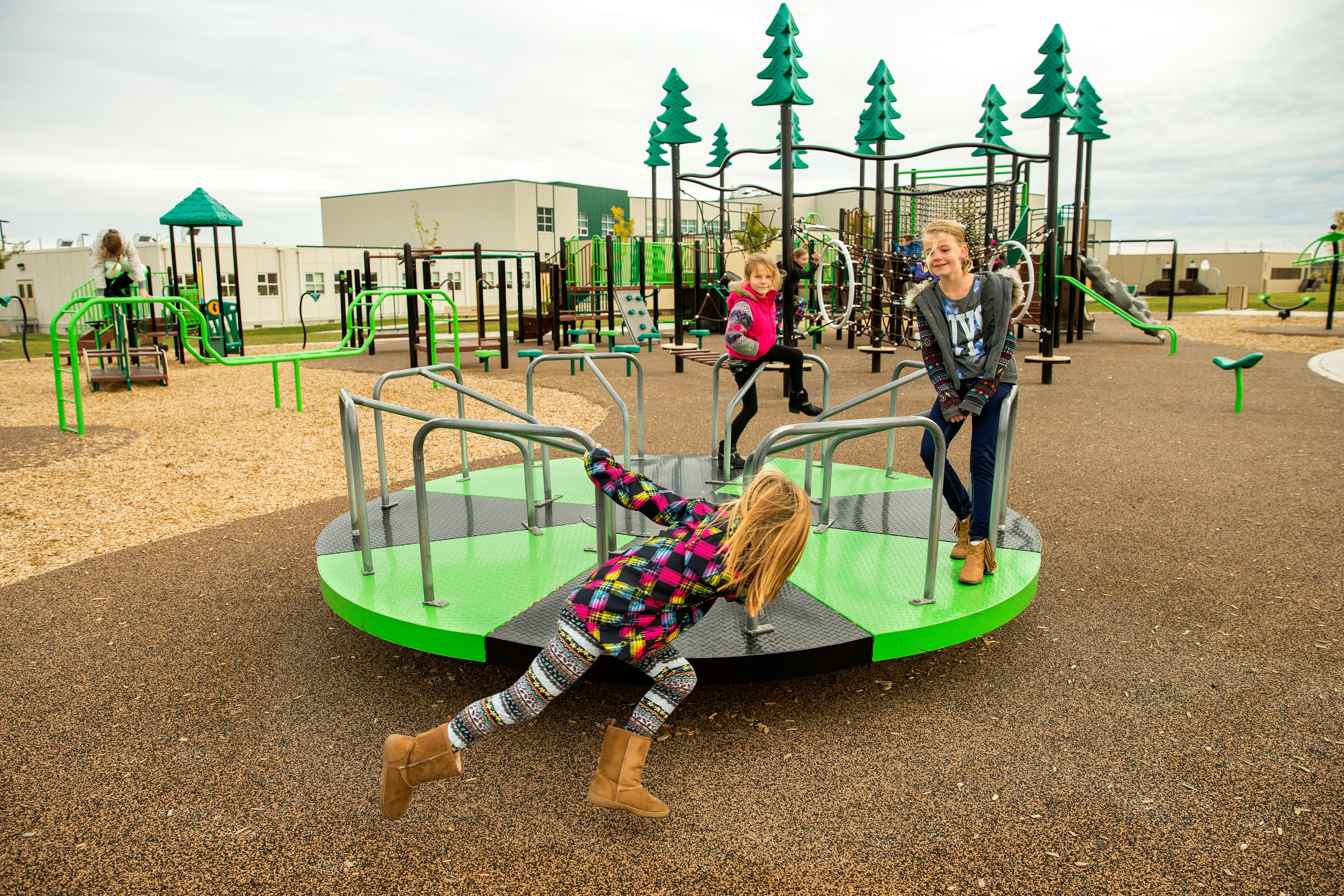 Children playing on a green-and-black merry-go-round in the foreground with climbing bars, balance elements and rope features behind.
