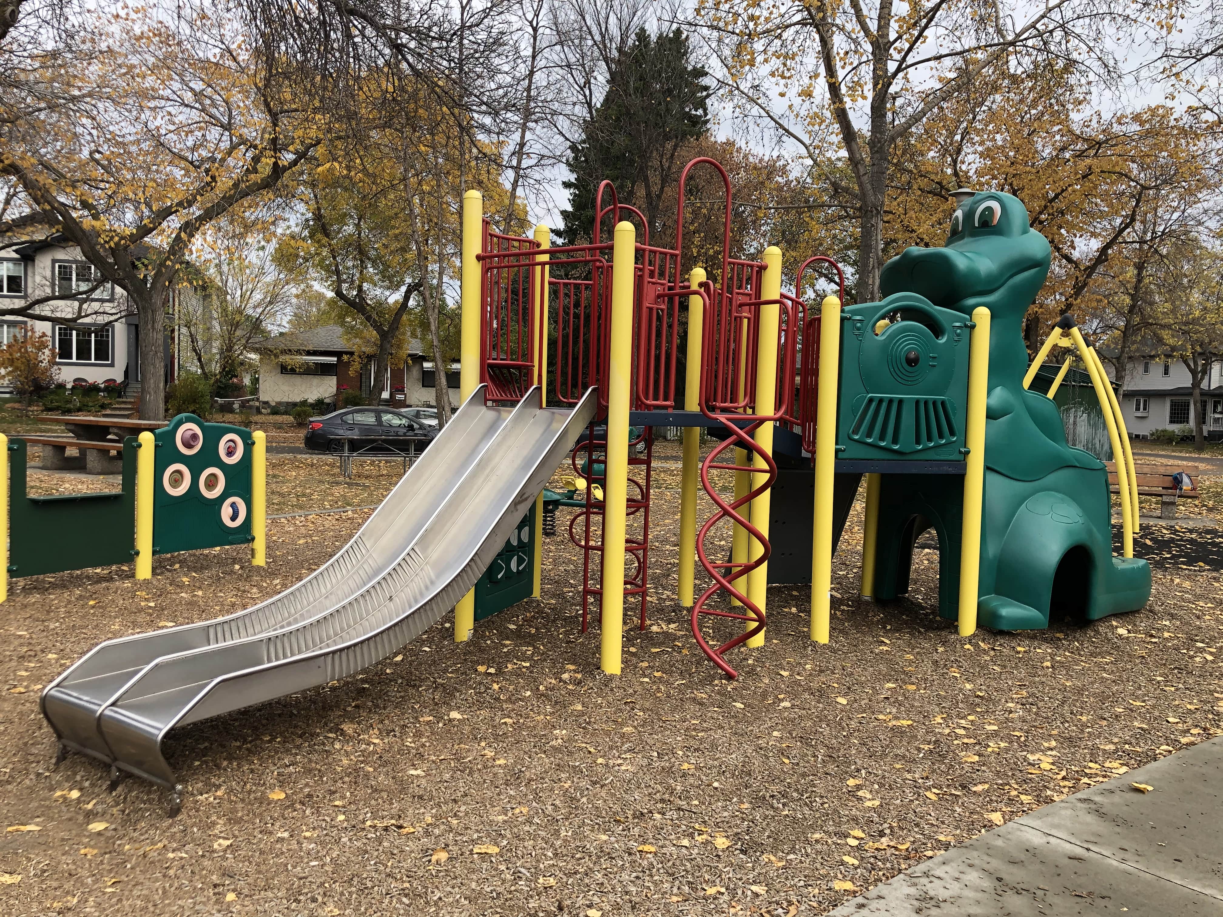 Yellow-and-red play structure with triple metal slides and a green smiling train-style slide feature, set against autumn trees.