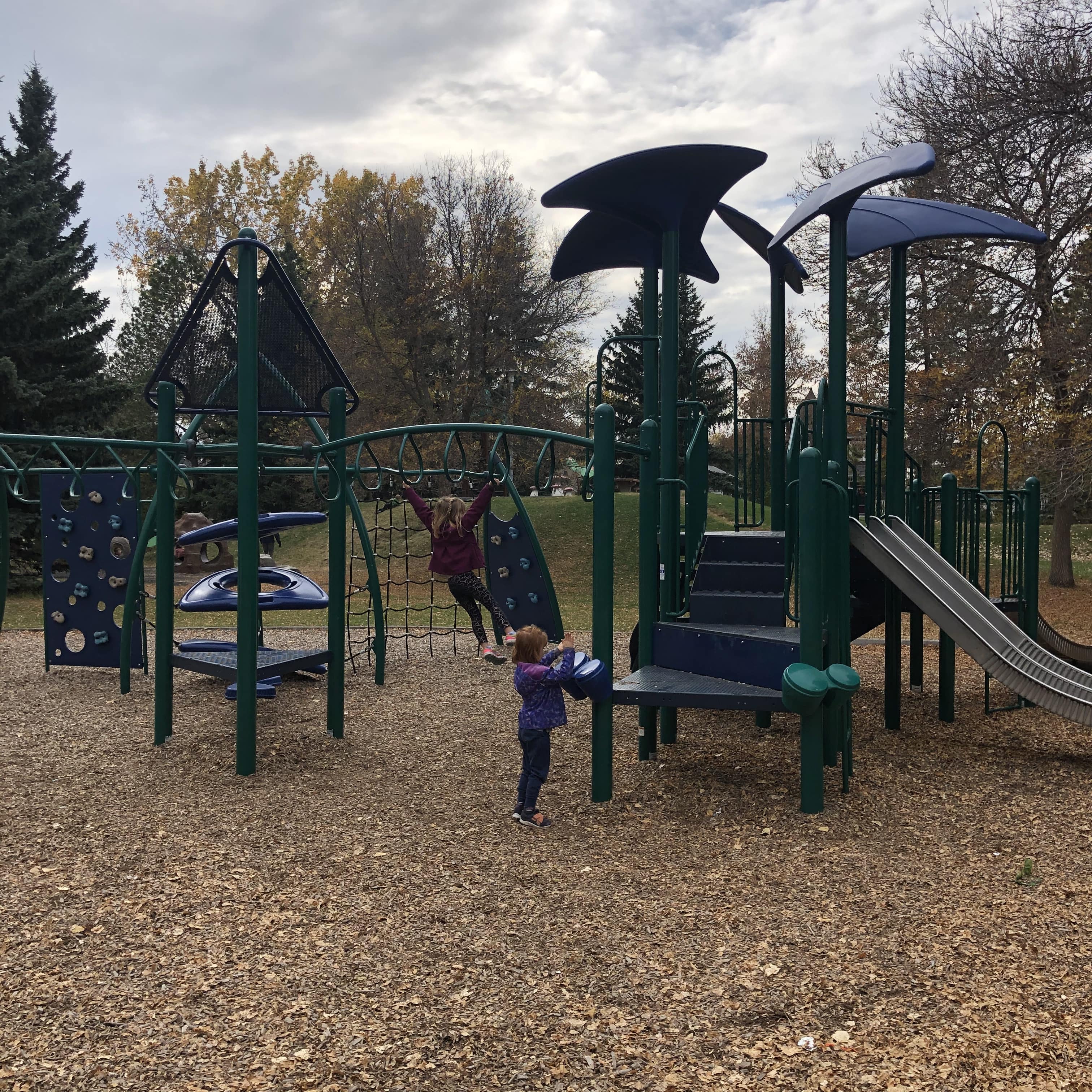 Large green playground structure with blue curved shade panels, climbing net and two children playing beneath.