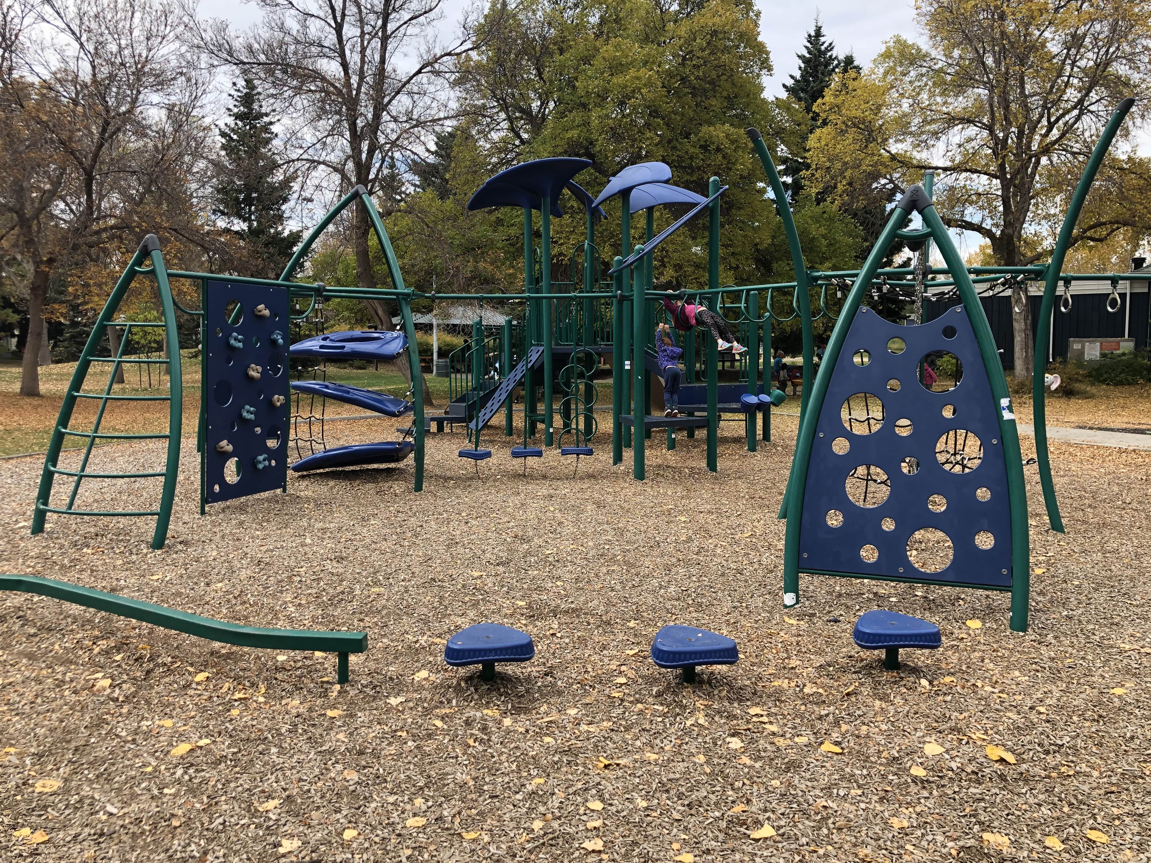 Green and blue playground structure with circular cutout panels, hanging rings and stepping pods on woodchip ground under trees.