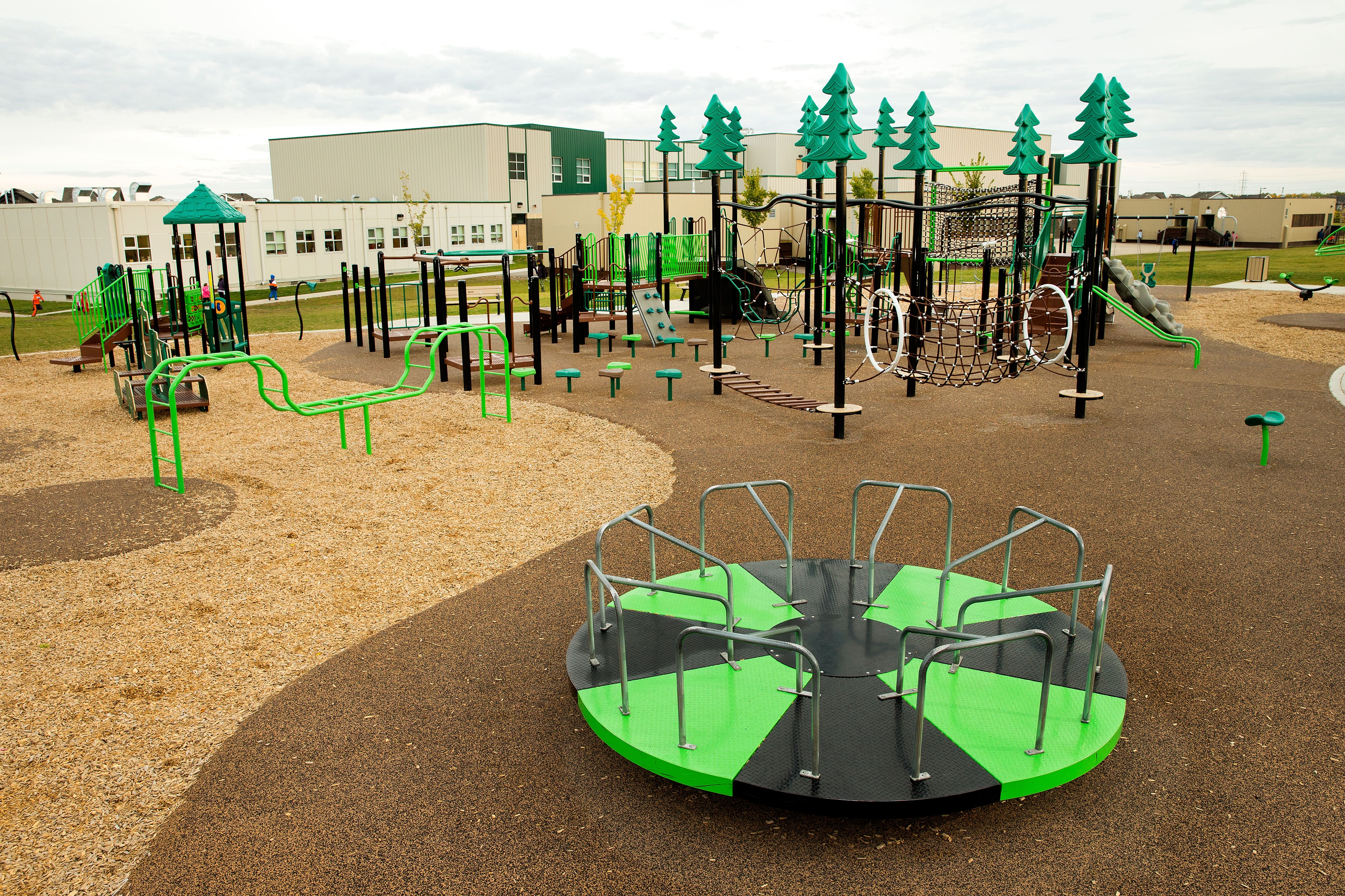 Wide view of the playground showing a green-and-black merry-go-round up front and an extensive rope-climb structure, slides and platforms beyond.