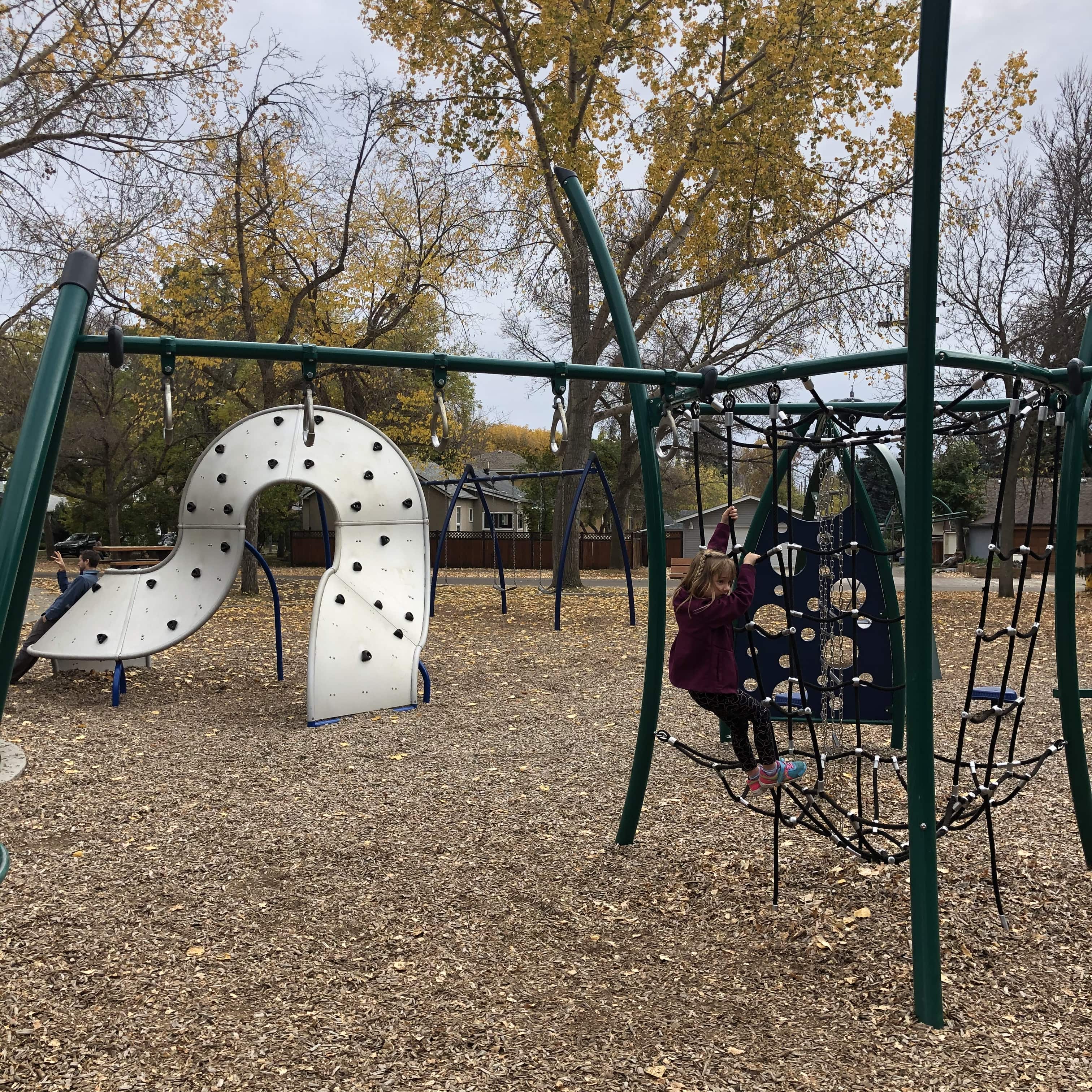 Horizontal monkey bars with hanging rings above a white curved climbing wall, fall trees behind and a child reaching up.
