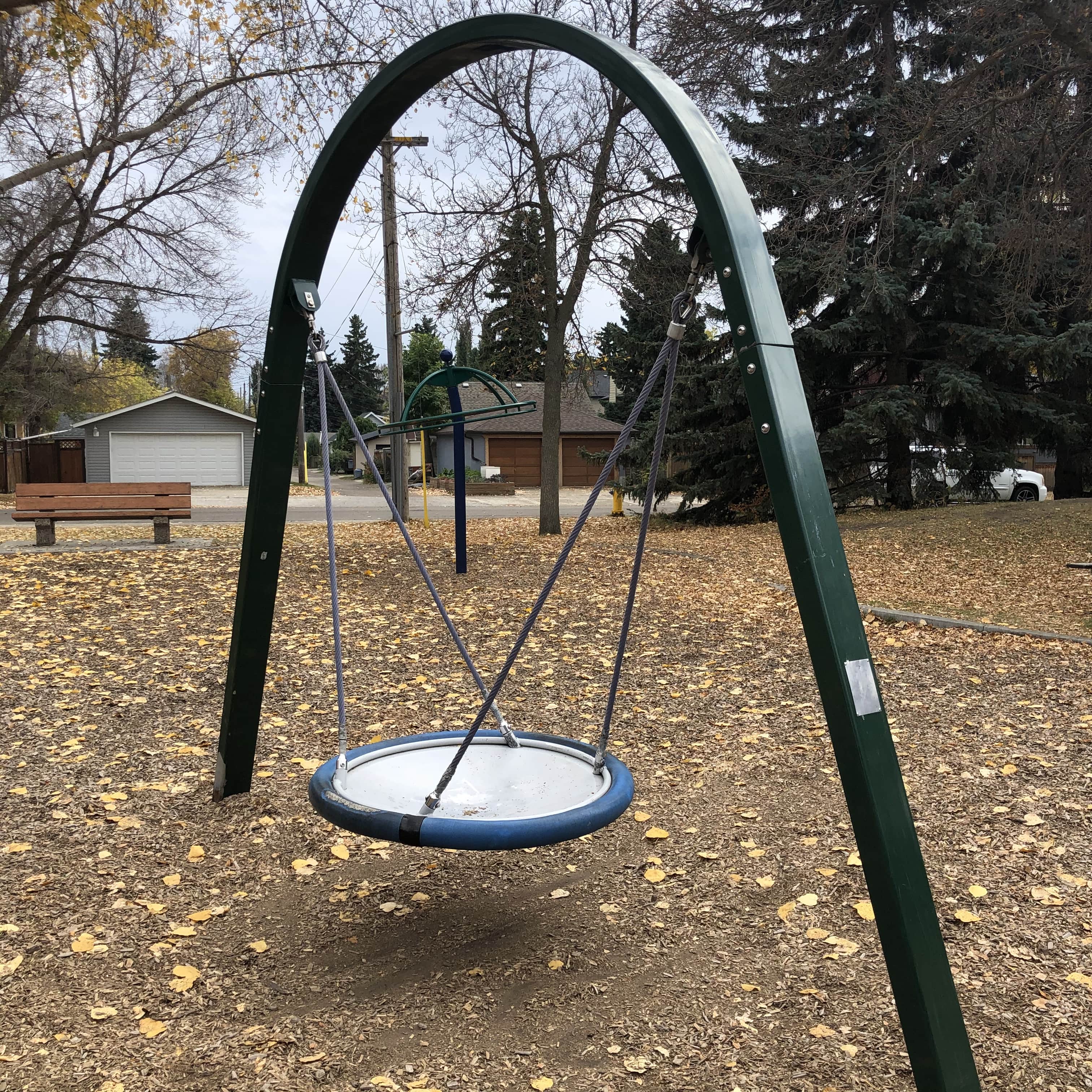 Round disk swing suspended by ropes from a green arch frame over wood-chip ground, with a bench and houses visible in the background.
