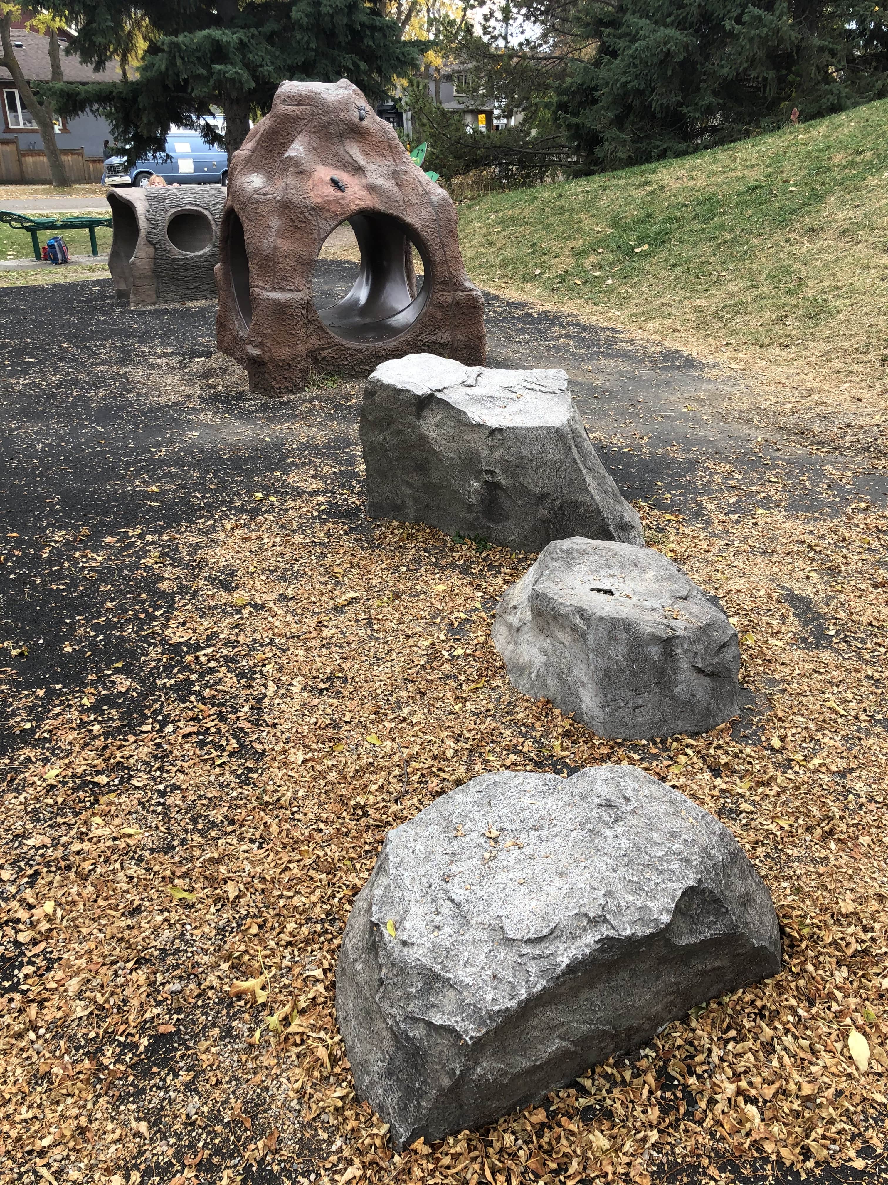 Brown rock-like tunnel play feature with large grey faux boulders on black rubber surfacing, scattered fall leaves around.